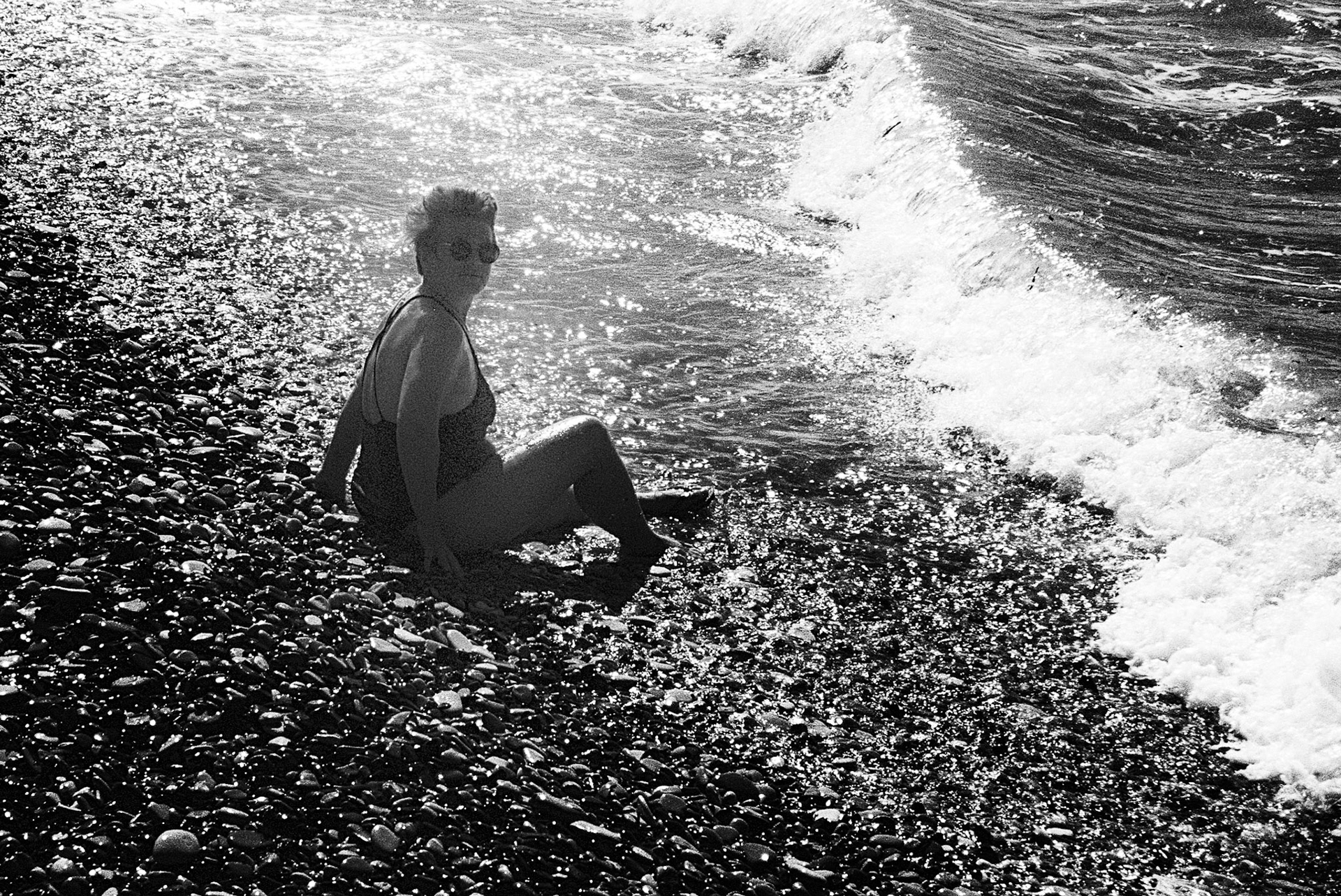 A lone figure sits where the waves meet the shore, sunlight scattering across water and stone.