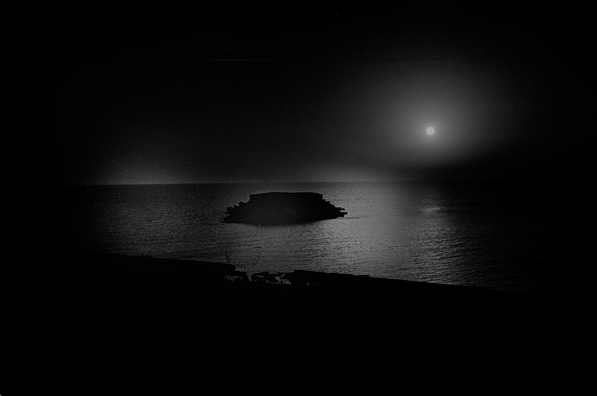A lone rocky outcrop sits in still water beneath a bright Mediterranean moon, its reflection shimmering across the bay.