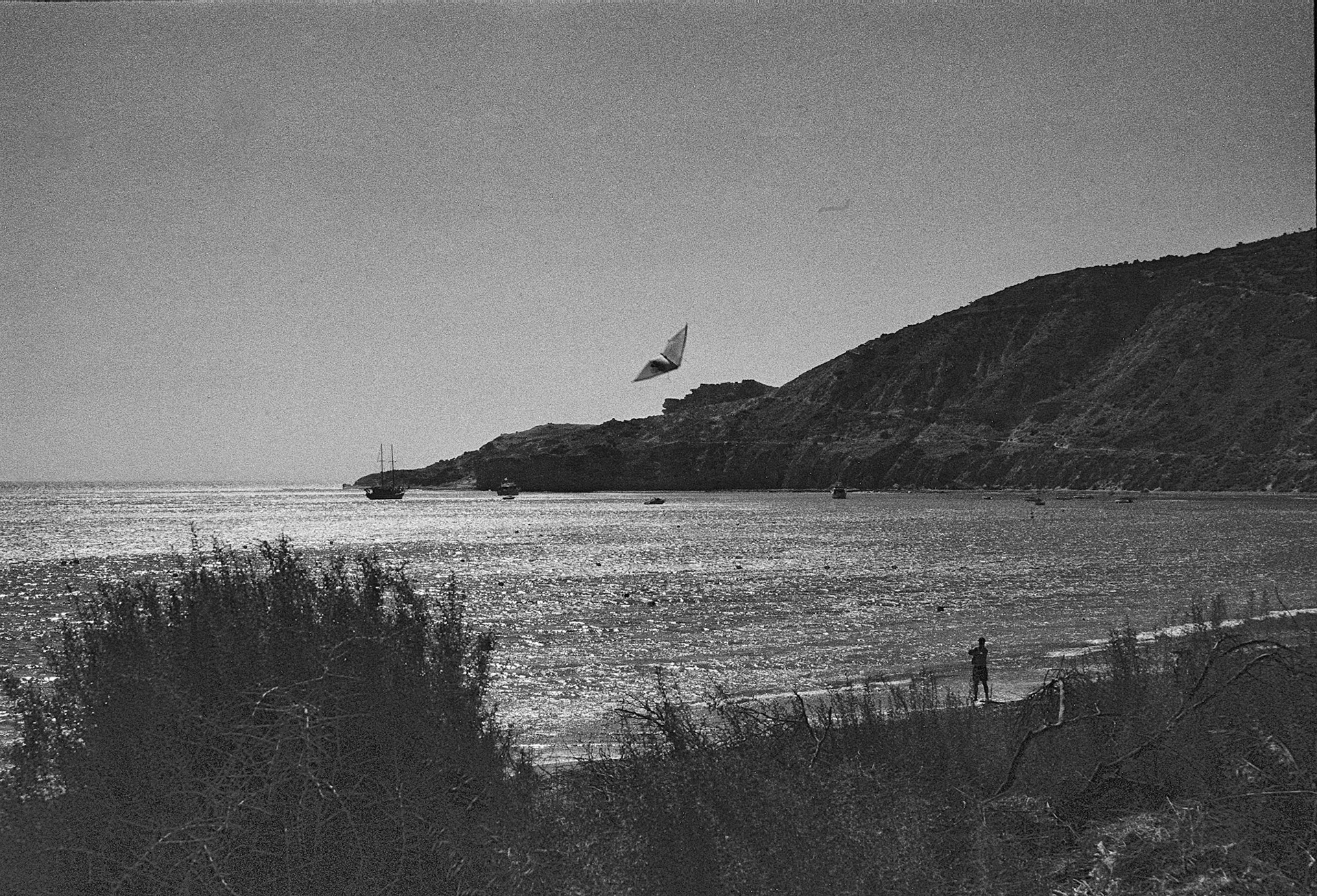 A lone kite drifts above a shimmering bay while a solitary figure watches the sea below.