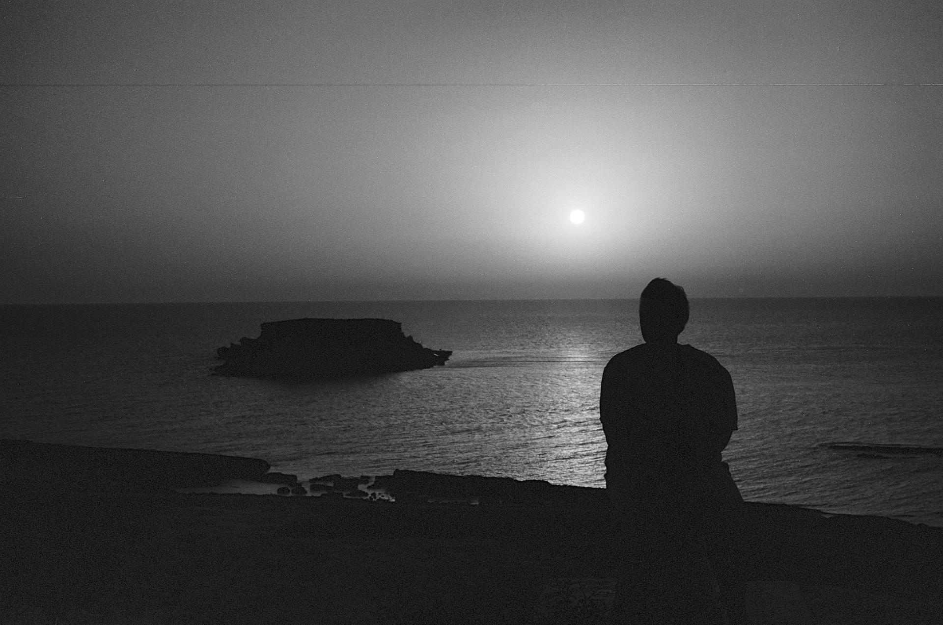 A solitary figure looks out across a calm Mediterranean sea as the sun (or moon) hangs low over a distant islet.