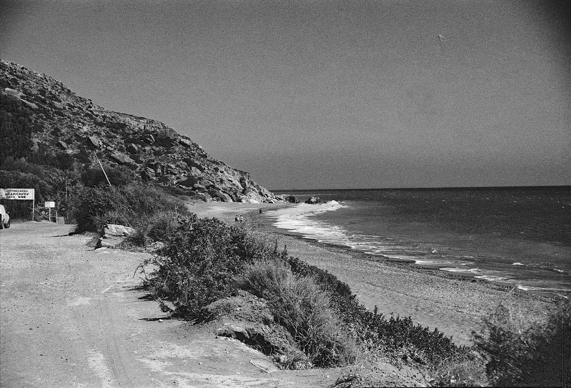 A dusty coastal road curves past scrub and rock toward a quiet pebble beach on the Mediterranean.