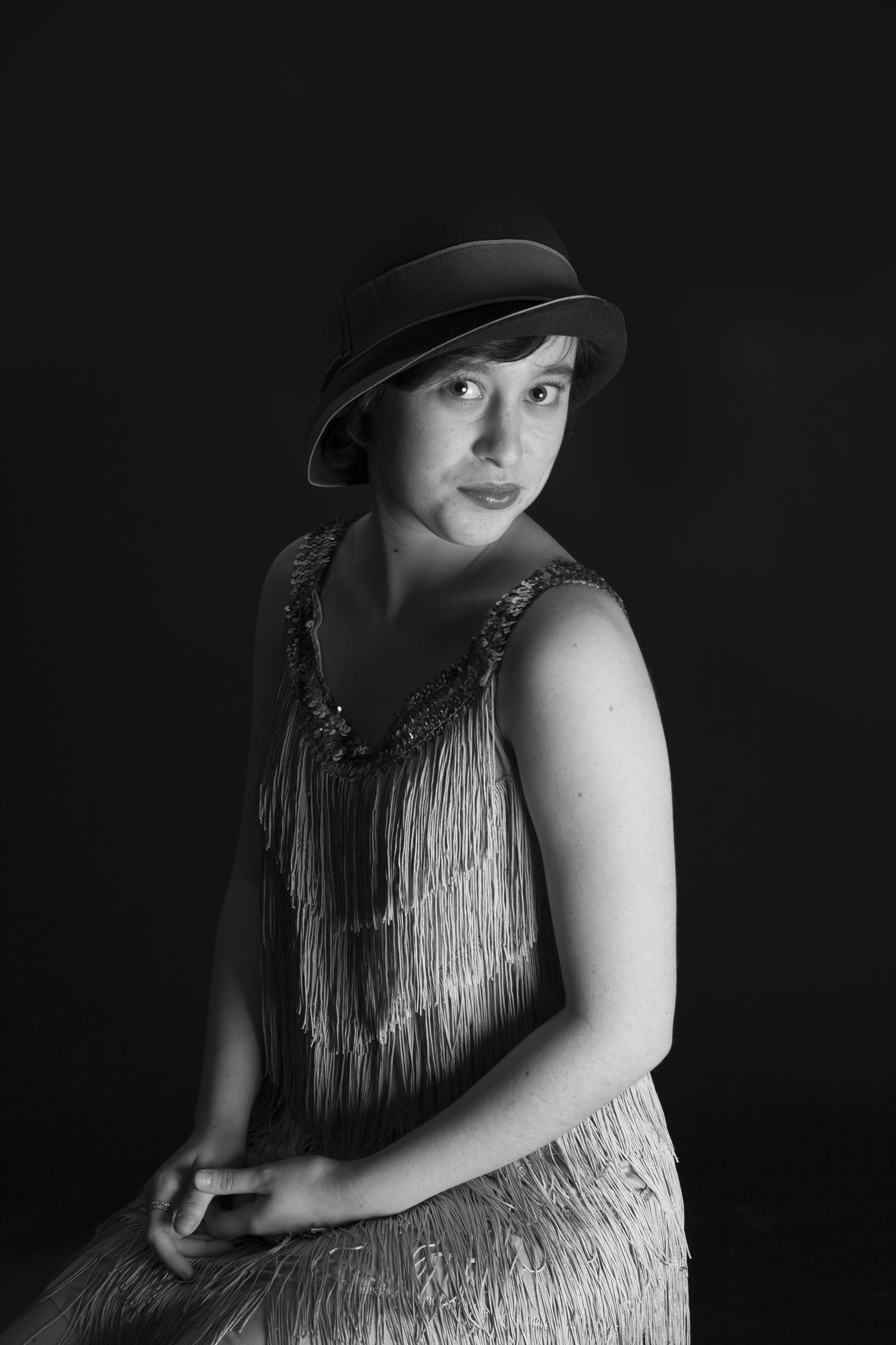 A studio portrait in black and white of a women dressed in a fringed 1920's flapper dress. She is wearing a cloche hat and wearing lipstick.