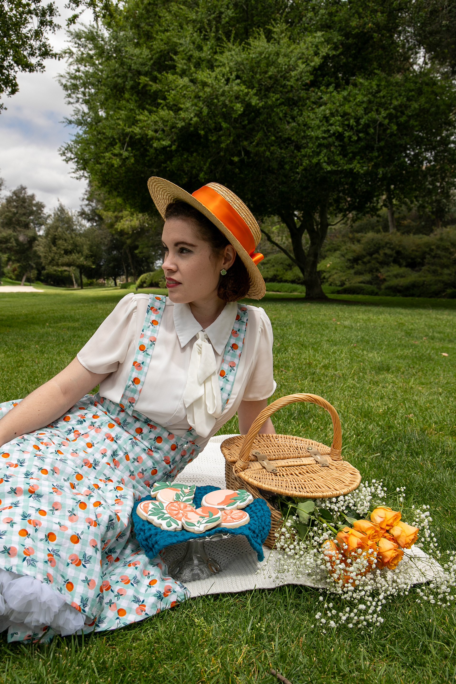 A vintage-inspired picnic scene featuring a woman in a floral dress and straw hat, surrounded by cookies and flowers.