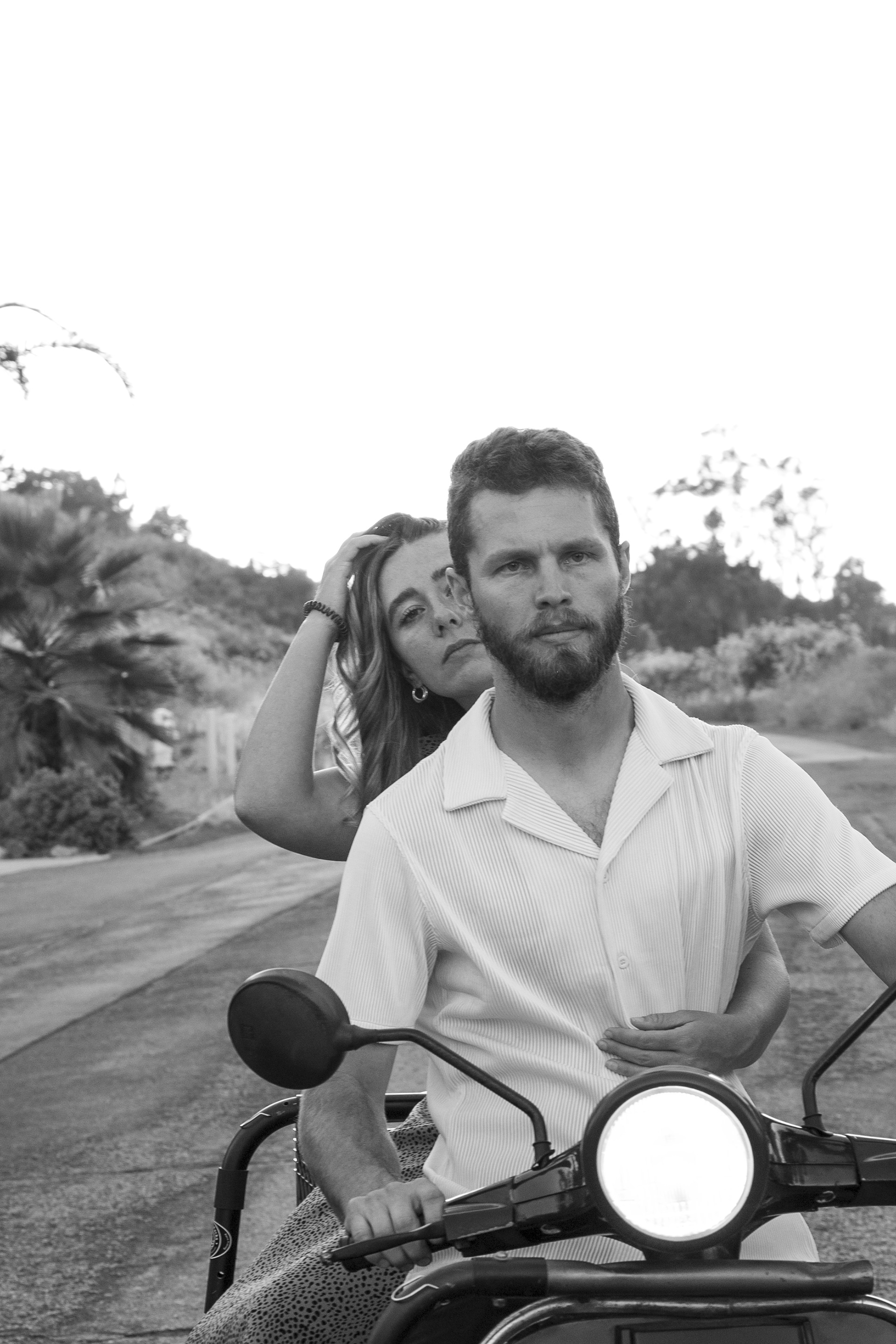 Image of a couple on a moped. This image is in black and white, girl is moving hair from face, man is facing camera with a concerned look. Both are wearing retro inspired cloths.