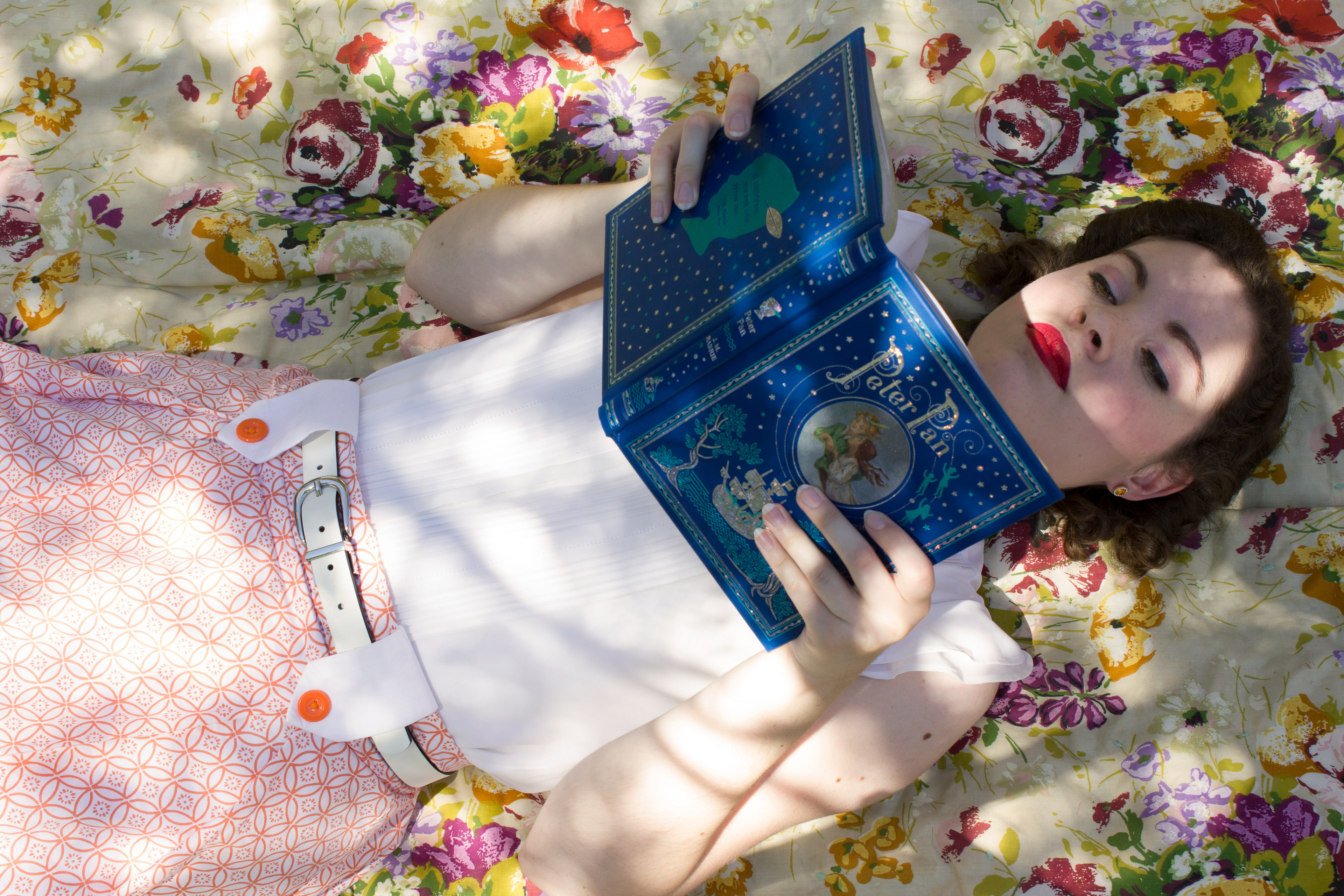 A women poses on a floral blanket reading Peter Pan in the shade of a tree. She is wearing a orange skirt and white belt that calls to the 1940's