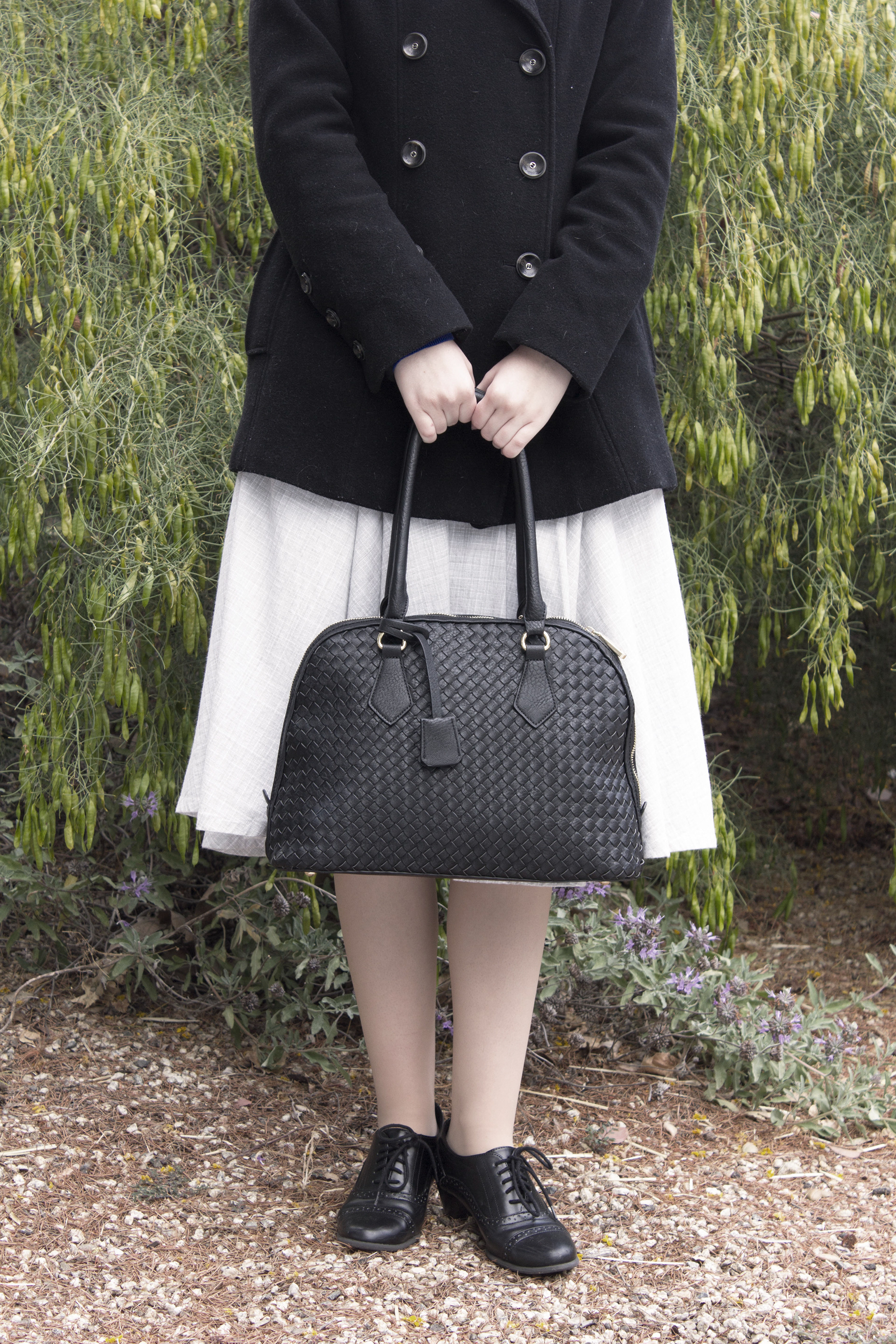 A portrait from the shoulders down focusing on this retro bag. There is symmetry between the black peacoat and the black purse that is Infront of the grey skirt. 