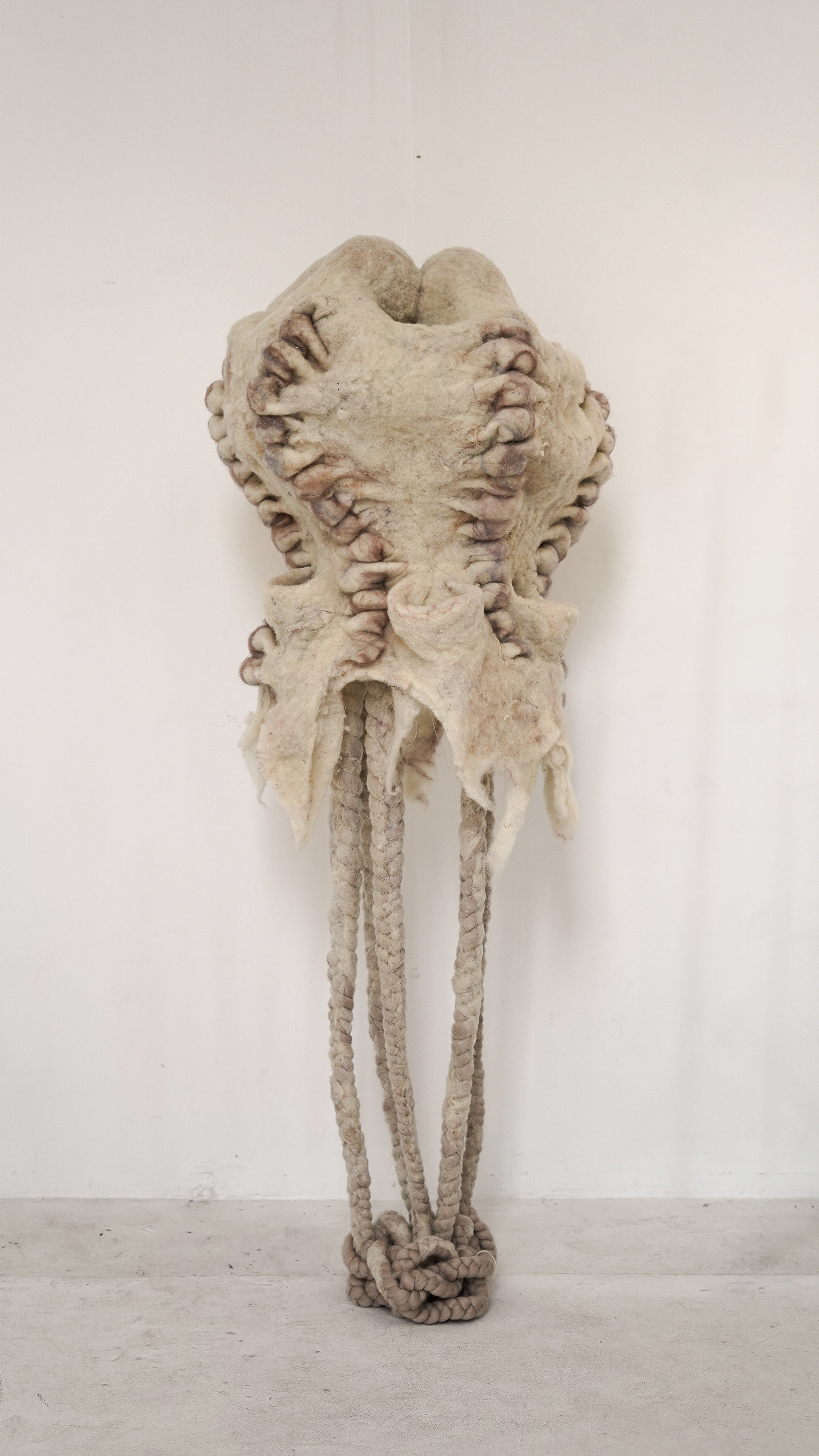 Hanging textile sculpture titled with us by Ludmila Bubanova. The artwork is made of light organic felted wool, resembling conjoined human figures with dark spiral seams on the sides and long, braided cords cascading to the floor.