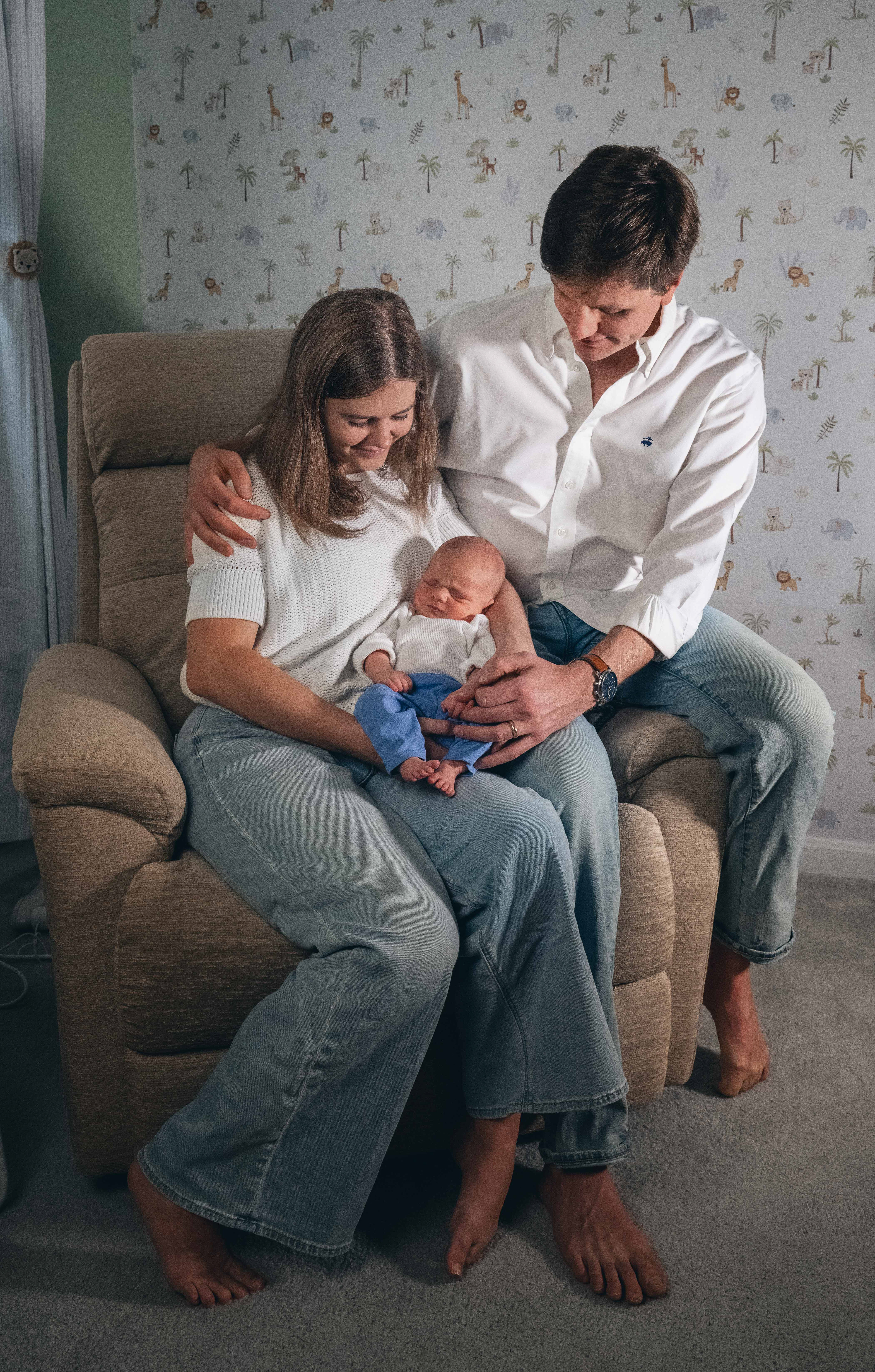 A man and a woman seated on a lounge chair looking down at their newborn baby.