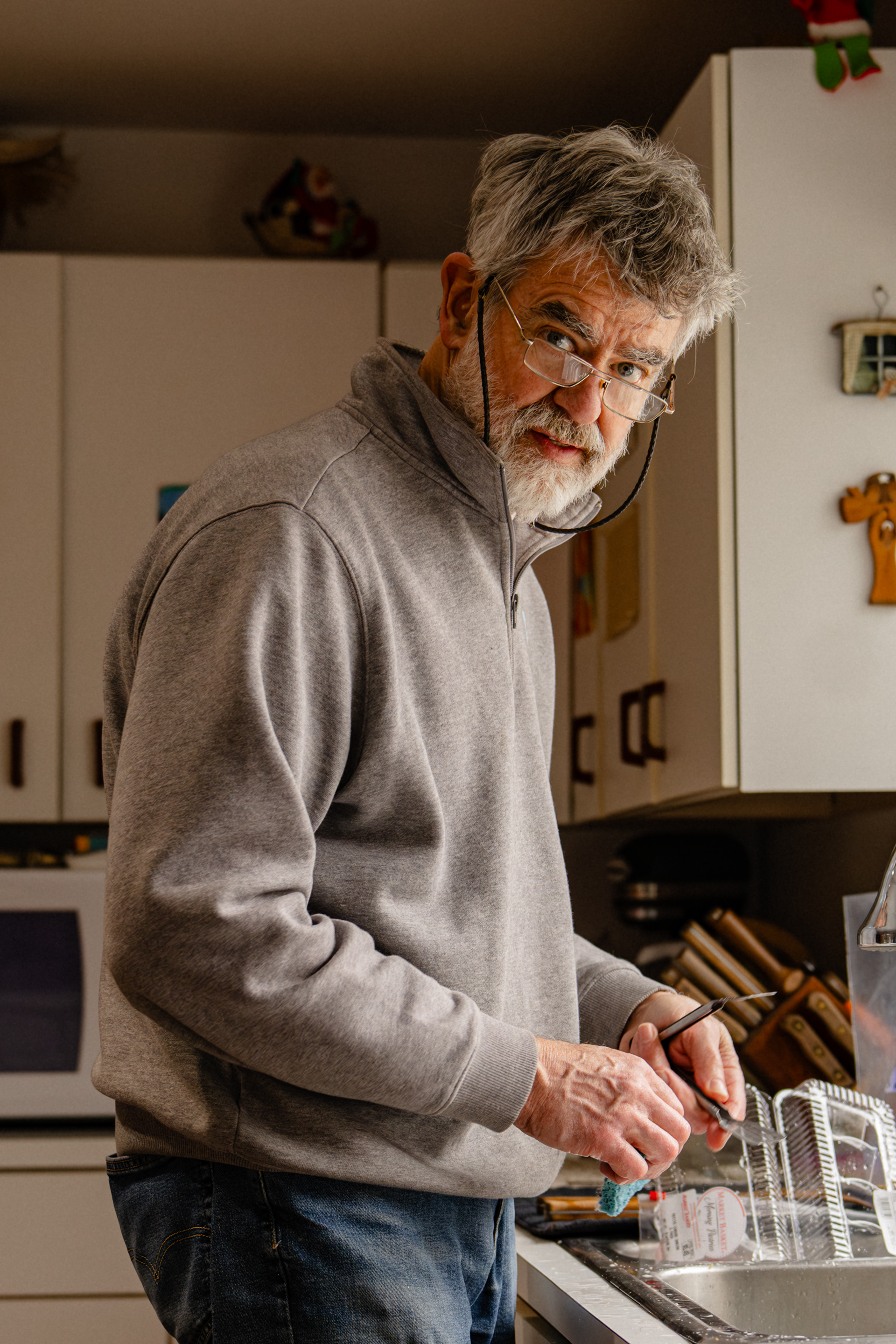 A man in his 60s with glasses looking at the camera while standing at the kitchen sink.