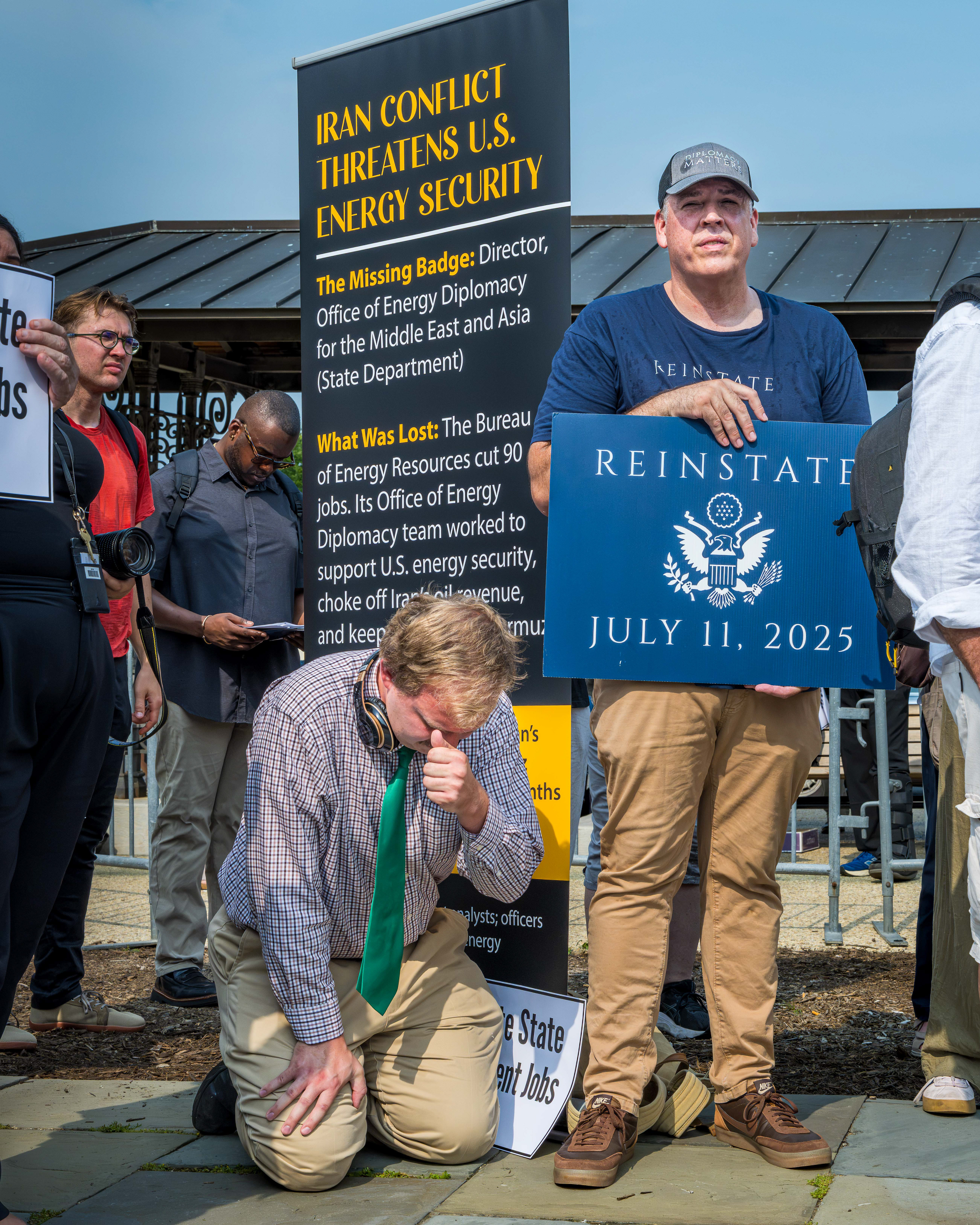 Two men, one kneeling, in from of signs about the Iran conflict and State Department firings.