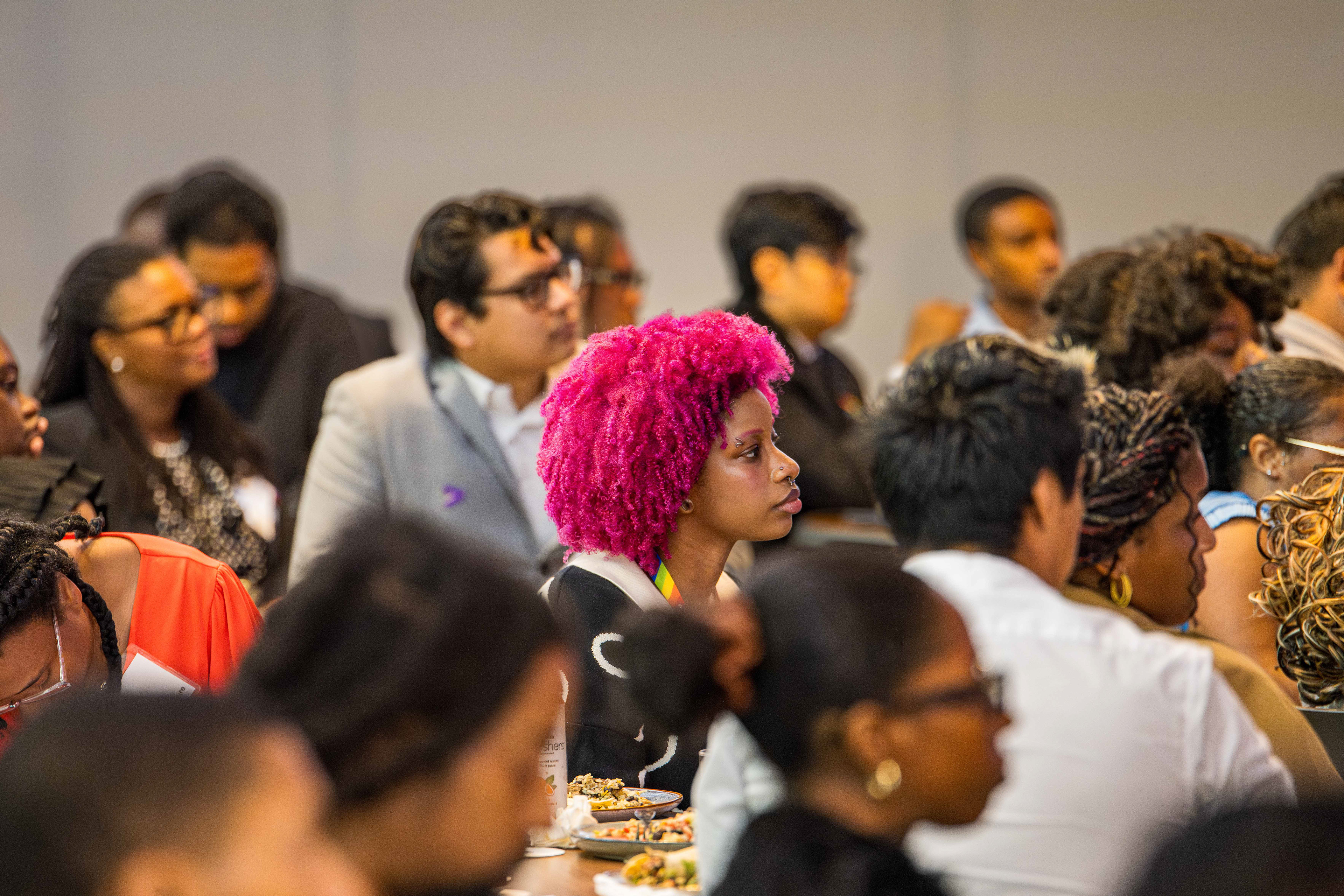 A Black woman with Pink hair in the middle of a conference looking at the presentation.