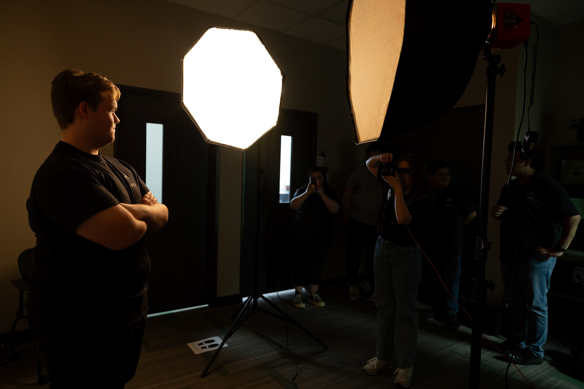 IN THE SPOTLIGHT- Sophomore Nathan Basset, poses for a headshot during the Feb. 4 2025 Hatch Visuals Studio Workshop. While behind the camera, Lauren Campbell, Managing Editor, explains camera settings and lighting techniques to the workshop attendees. Photo By Alaina Jones