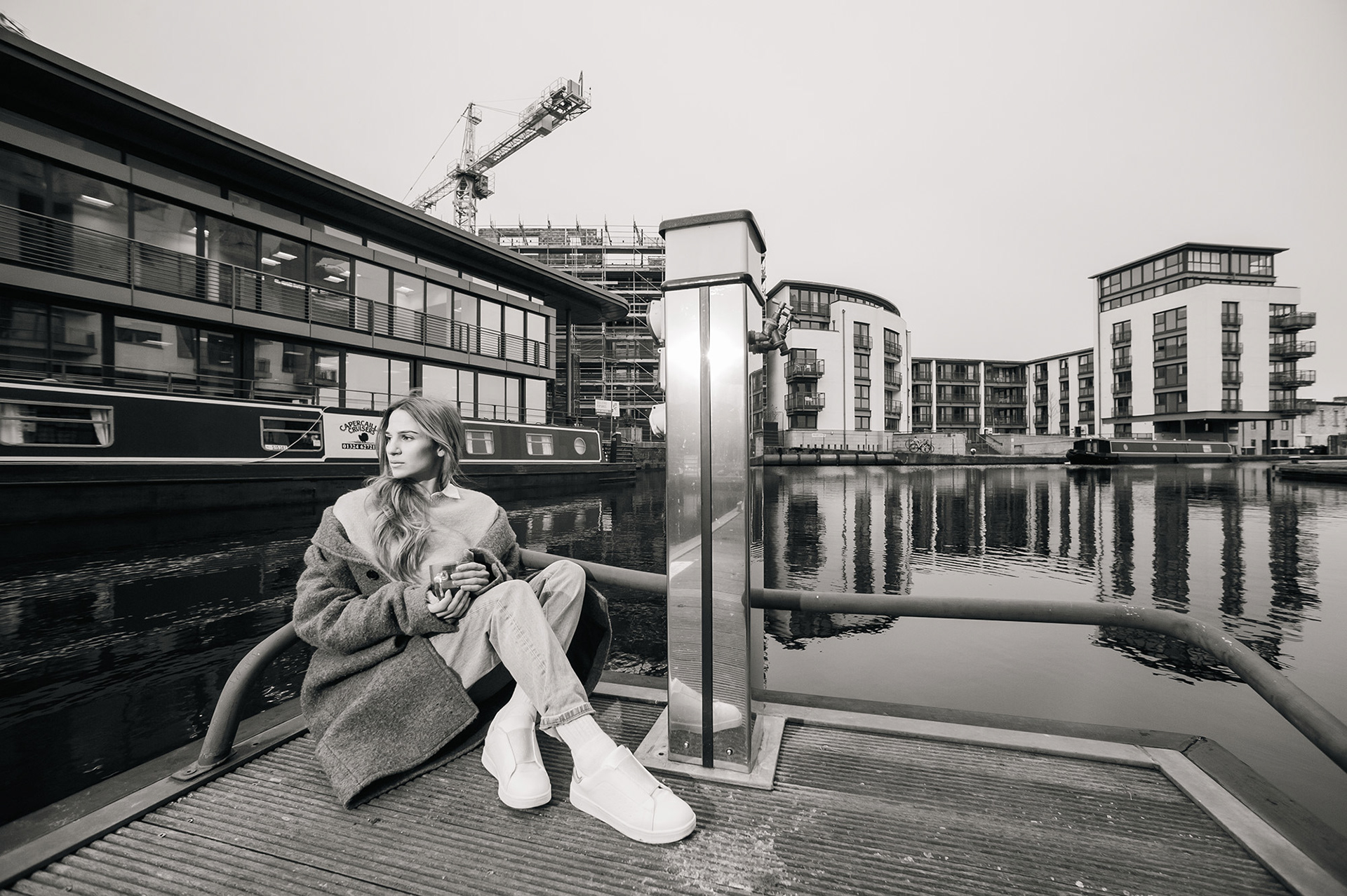 Γυναίκα κάθεται δίπλα σε κανάλι με φόντο σύγχρονα κτίρια και γερανούς, ντυμένη σε casual minimal στυλ.    Woman sitting by a canal with modern buildings and cranes in the background, dressed in casual minimal style.