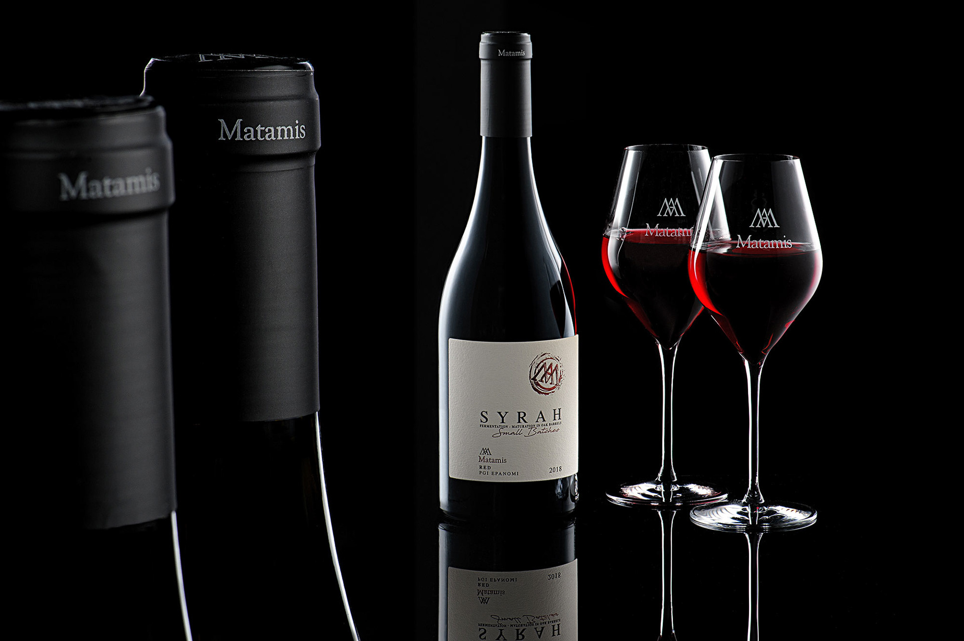 διαφημιστική φωτογραφία κόκκινου κρασιού syrah matamis wine μπουκάλι με 2 ποτήρια γεμάτα σε μάυρο φόντο με αντανάκλαση