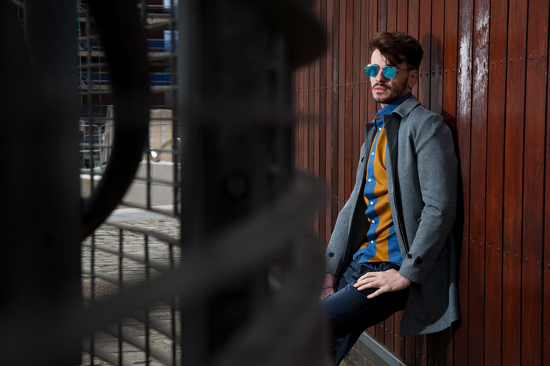 Άνδρας με γκρι παλτό και πουκάμισο σε έντονα χρώματα ποζάρει δίπλα σε ξύλινο τοίχο σε αστικό περιβάλλον.,       Man in grey coat and colorful shirt leaning against a wooden wall in an urban location.    