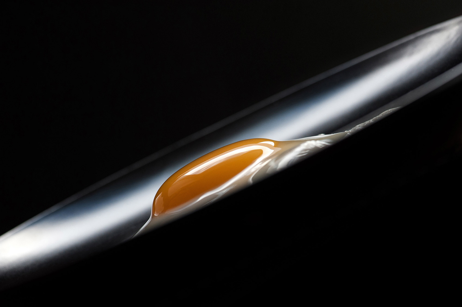 Μια μινιμαλιστική φωτογραφία fine art food photography με έναν λαμπερό, ρευστό κρόκο αβγού που ισορροπεί αριστοτεχνικά πάνω σε μια κομψή, μεταλλική καμπύλη με έντονες αντανακλάσεις σε μαύρο φόντο, από τη φωτογράφο Βούλα Γκώτη. A minimalist fine art food photography capturing a glossy, fluid egg yolk perfectly balanced on a sleek metallic curve with sharp reflections against a dark background, by photographer Voula Gkoti.
