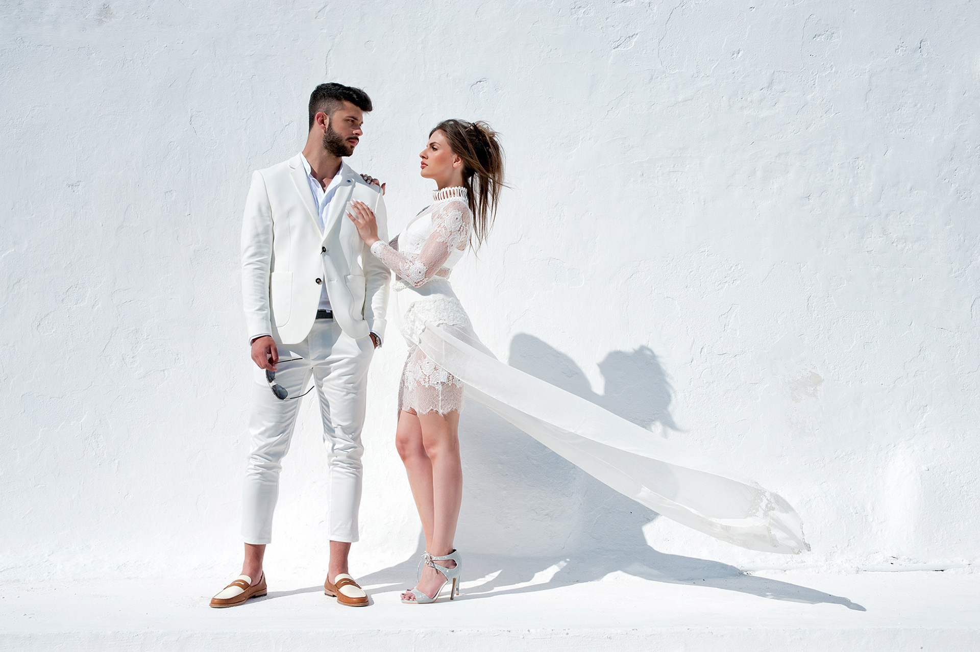 Ζευγάρι ντυμένο στα λευκά ποζάρει μπροστά σε λευκό τοίχο με καθαρές γραμμές και απλότητα.,         Elegant fashion couple dressed in white posing together against a minimalist white wall.    