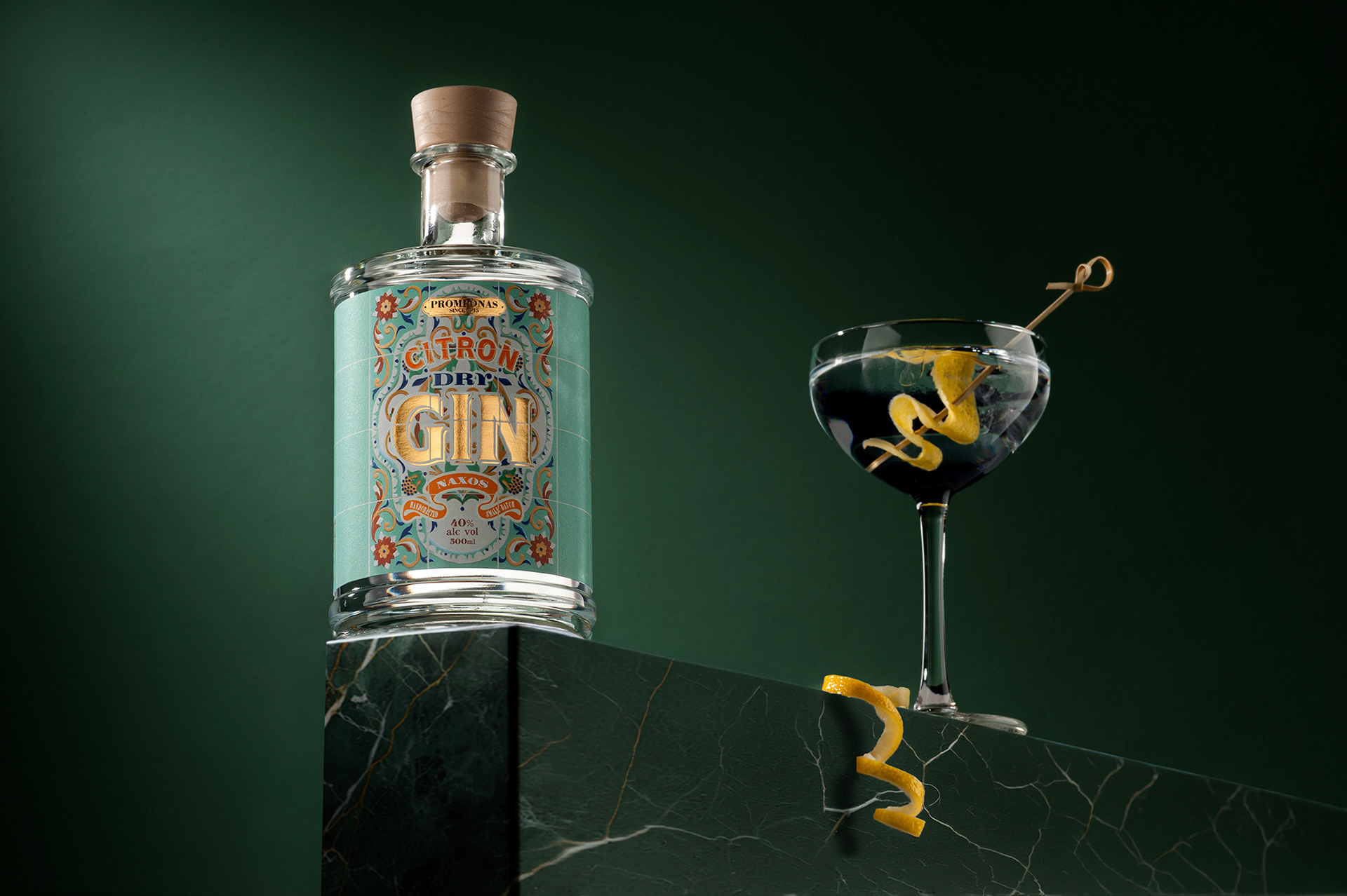 Φωτογράφιση μπουκάλι Citron Dry Gin πάνω σε μαρμάρινη βάση δίπλα σε cocktail martini με φλούδα λεμονιού, σε σκούρο πράσινο φόντο,   Bottle of Citron Dry Gin on a marble pedestal next to a martini cocktail with a lemon twist, set against a dark green background