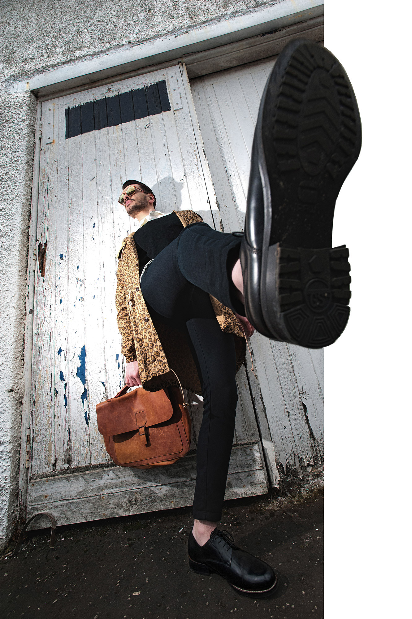 Άνδρας με λεοπάρ παλτό και δερμάτινη τσάντα ποζάρει δυναμικά με παπούτσι στο προσκήνιο.  Man wearing a leopard coat and leather bag posing dynamically with his shoe in the foreground.
