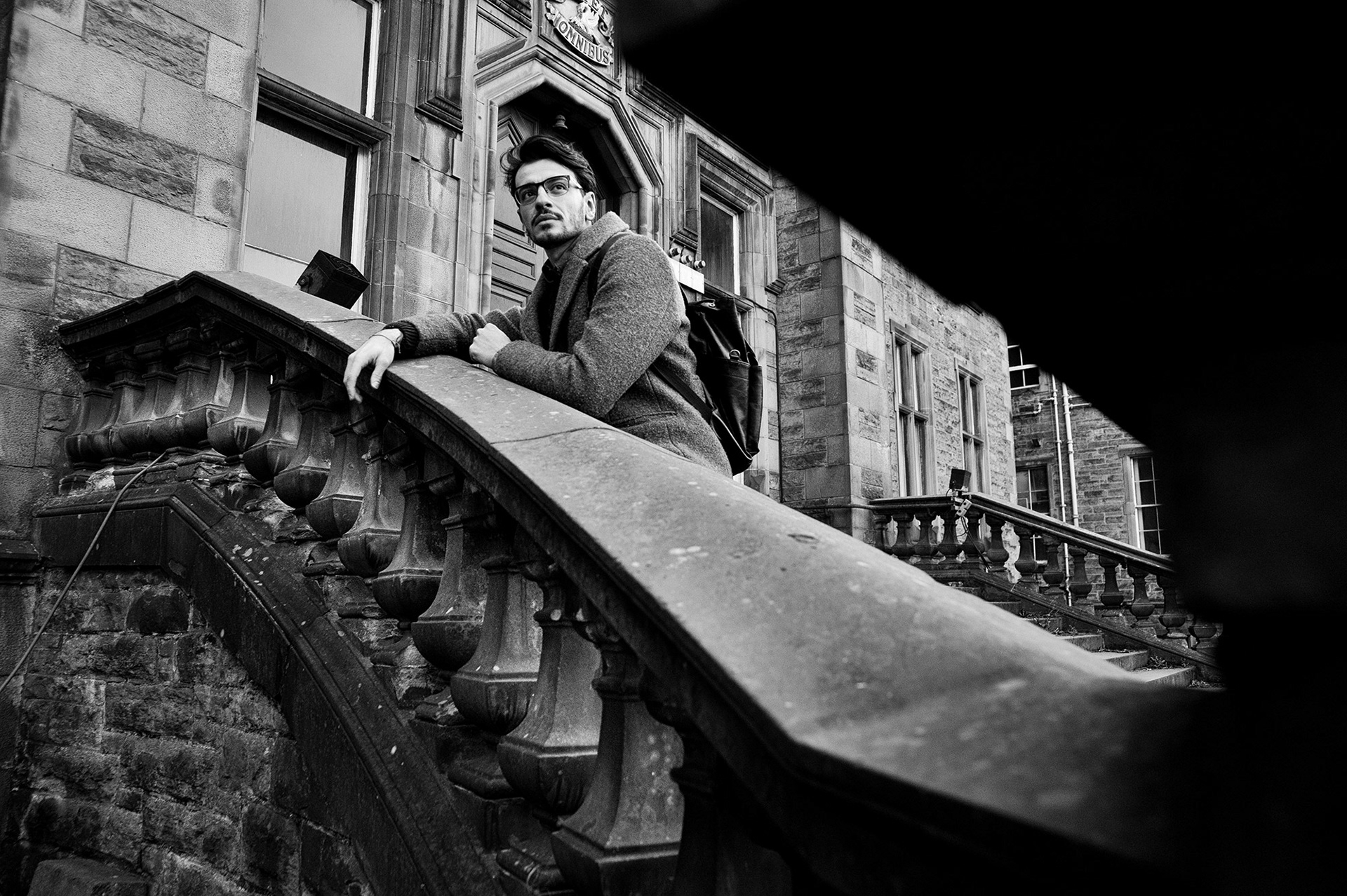 Άνδρας σε γκρι παλτό στέκεται σε πέτρινη σκάλα παλιού κτιρίου, κοιτάζοντας μακριά.    Man in a grey coat standing on stone stairs of an old building, looking into the distance.