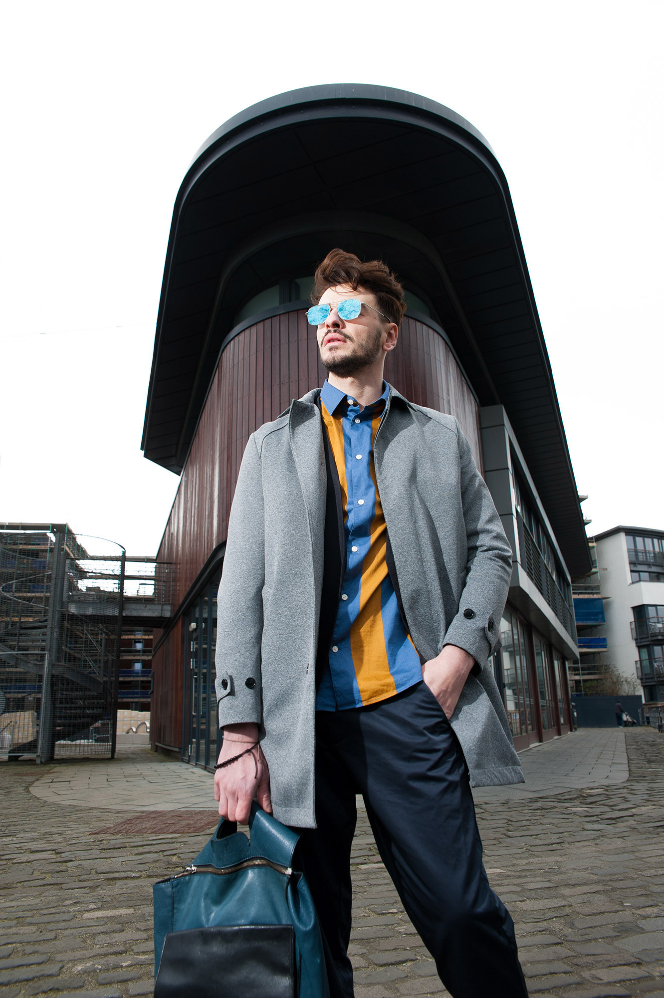 Άνδρας με γκρι παλτό και γυαλιά καθρέφτες ποζάρει μπροστά σε μοντέρνο κτίριο.            Man wearing a grey coat and mirrored sunglasses posing in front of a modern architectural building.