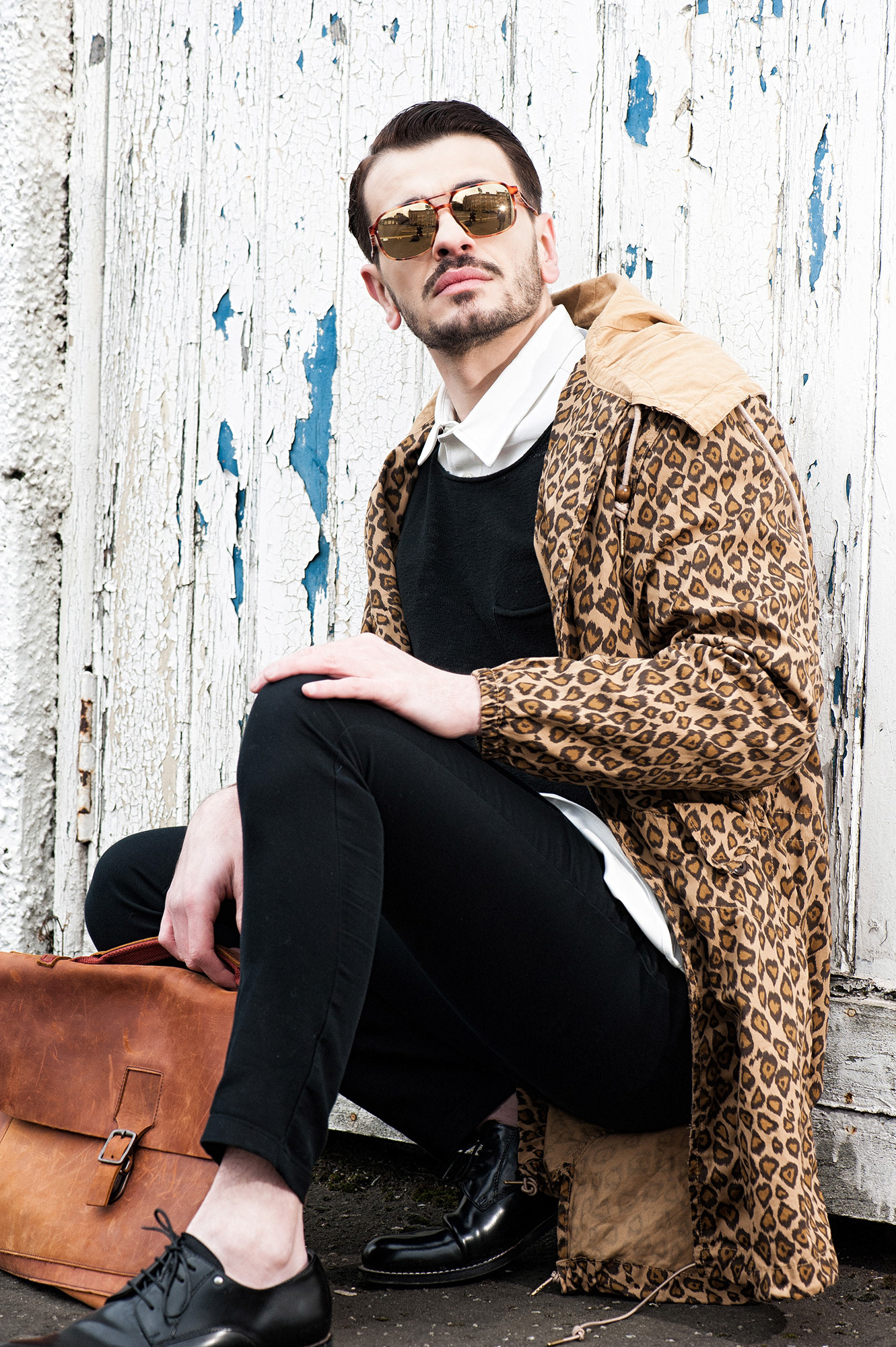Άνδρας με λεοπάρ παλτό, μαύρο πουκάμισο και δερμάτινη τσάντα κάθεται μπροστά σε παλιά ξύλινη πόρτα με ξεφτισμένη μπογιά.     Man wearing a leopard-print coat, black shirt, and leather bag sitting in front of an old weathered wooden door.