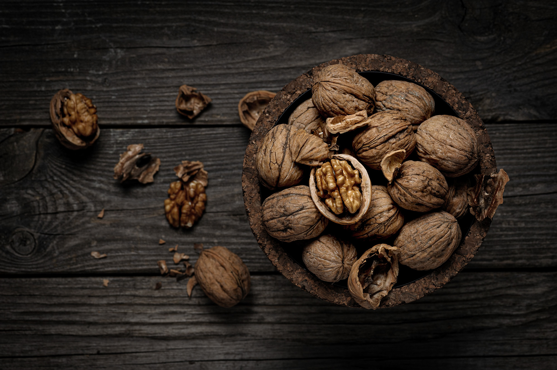 Μια ρουστίκ φωτογραφία fine art food photography με ολόκληρα καρύδια μέσα σε ένα χειροποίητο μπολ από φελλό, τοποθετημένα πάνω σε μια σκούρα, ανάγλυφη ξύλινη επιφάνεια, από τη φωτογράφο Βούλα Γκώτη.  A rustic fine art food photography shot featuring whole walnuts in a handmade cork bowl, arranged on a dark weathered wooden background, by photographer Voula Gkoti.