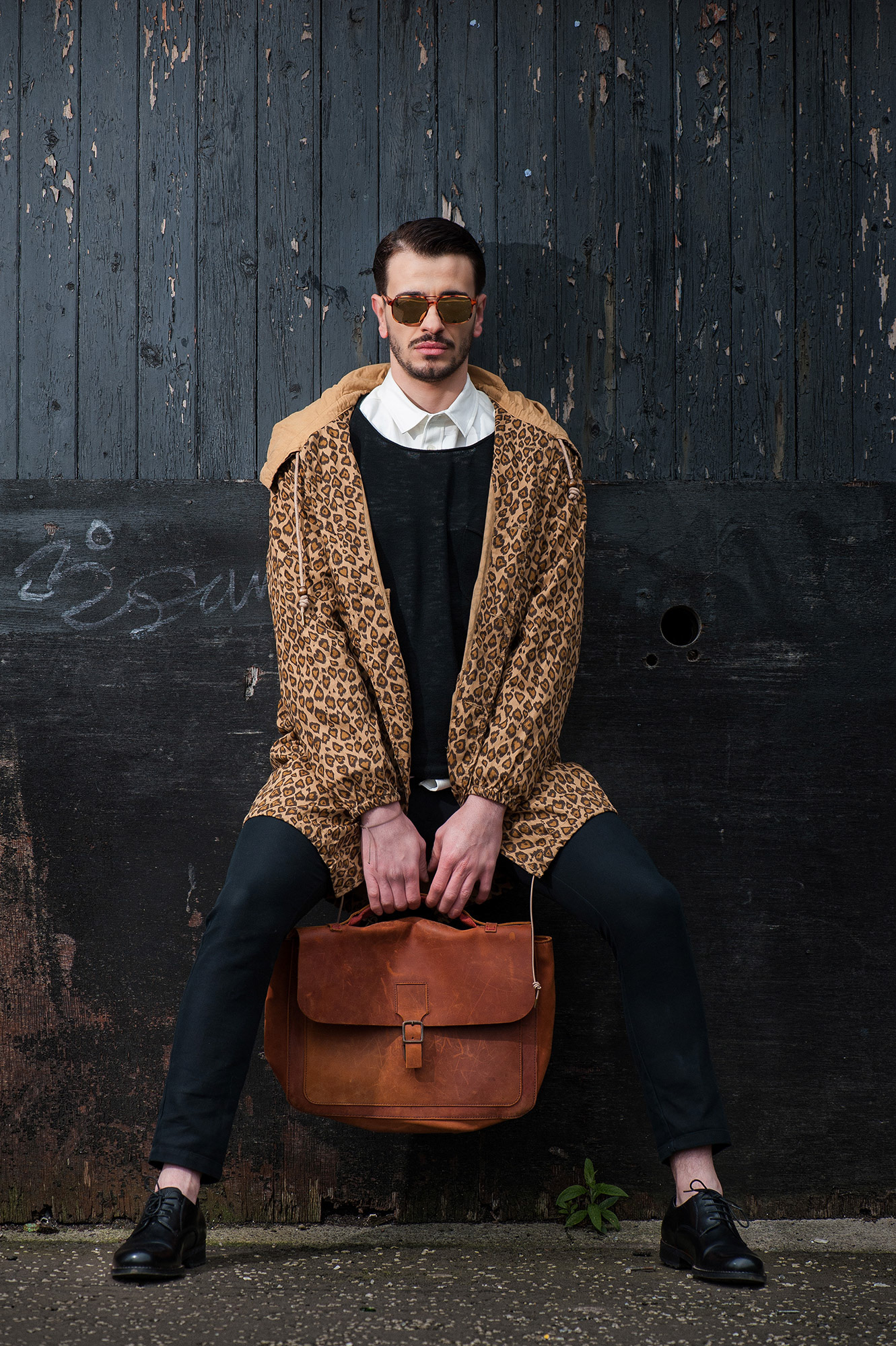 Άνδρας με λεοπάρ παλτό και γυαλιά καθρέφτες κάθεται μπροστά σε μαύρο ξύλινο φόντο κρατώντας καφέ τσάντα.      Man in a leopard jacket and mirrored sunglasses sits against a dark wooden wall holding a brown leather bag.