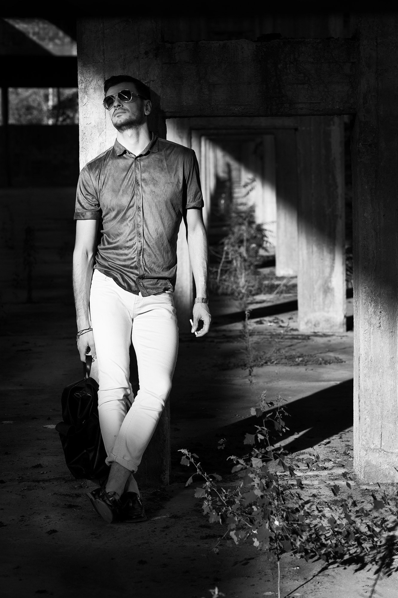 Άνδρας με γυαλιά ηλίου και ανοιχτό πουκάμισο ακουμπά σε τσιμεντένια κολόνα μέσα σε βιομηχανικό χώρο.    Man wearing sunglasses and a casual shirt leans against a concrete column in an industrial setting.