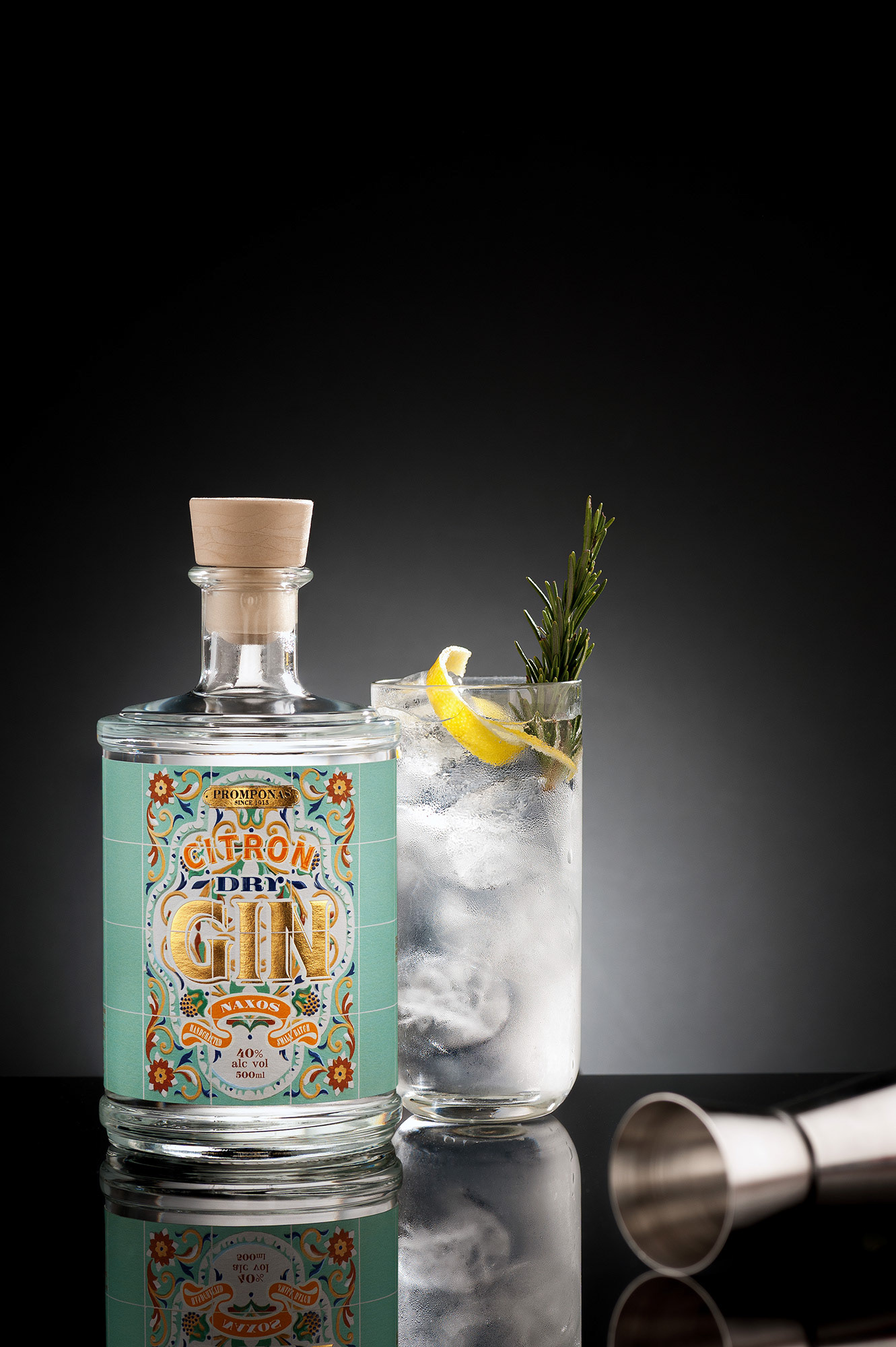 Μπουκάλι Citron Dry Gin σε μαύρο φόντο δίπλα σε ποτήρι gin tonic με πάγο, φλούδα λεμονιού και κλωνάρι δεντρολίβανου, πάνω σε γυαλιστερή επιφάνεια με μεταλλικό jigger στο προσκήνιο,   Bottle of Citron Dry Gin on a black background next to a gin and tonic glass with ice, lemon peel, and a rosemary sprig, placed on a glossy reflective surface with a metal jigger in the foreground
