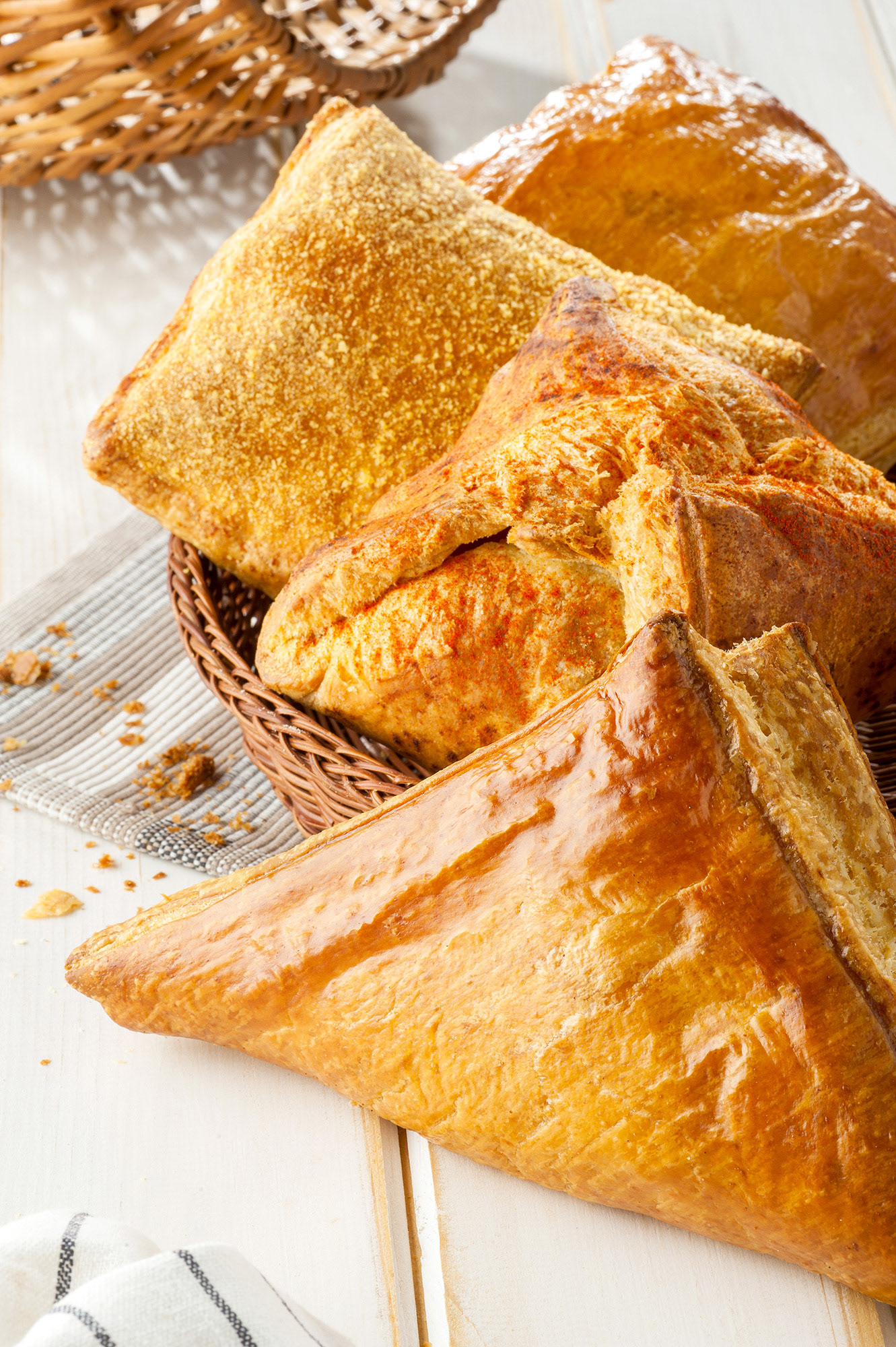 Φωτεινό food styling με μια ποικιλία από σφολιάτες μέσα σε ψάθινο καλάθι, που περιλαμβάνει τρίγωνη τυρόπιτα, πιτάκι με πάπρικα και γέμιση πίτσας, παραλληλόγραμμη σφολιάτα με γραβιέρα Νάξου και πίτες με τυρί κρέμα, αναδεικνύοντας τις τραγανές στρώσεις τους   /     Bright food styling of assorted puff pastry pies in a wicker basket, featuring a triangular cheese pie, a paprika-spiced pastry with pizza filling, a rectangular turnover stuffed with authentic Naxos gruyere cheese, and pastries filled with rich cream cheese, showcasing their flaky crust