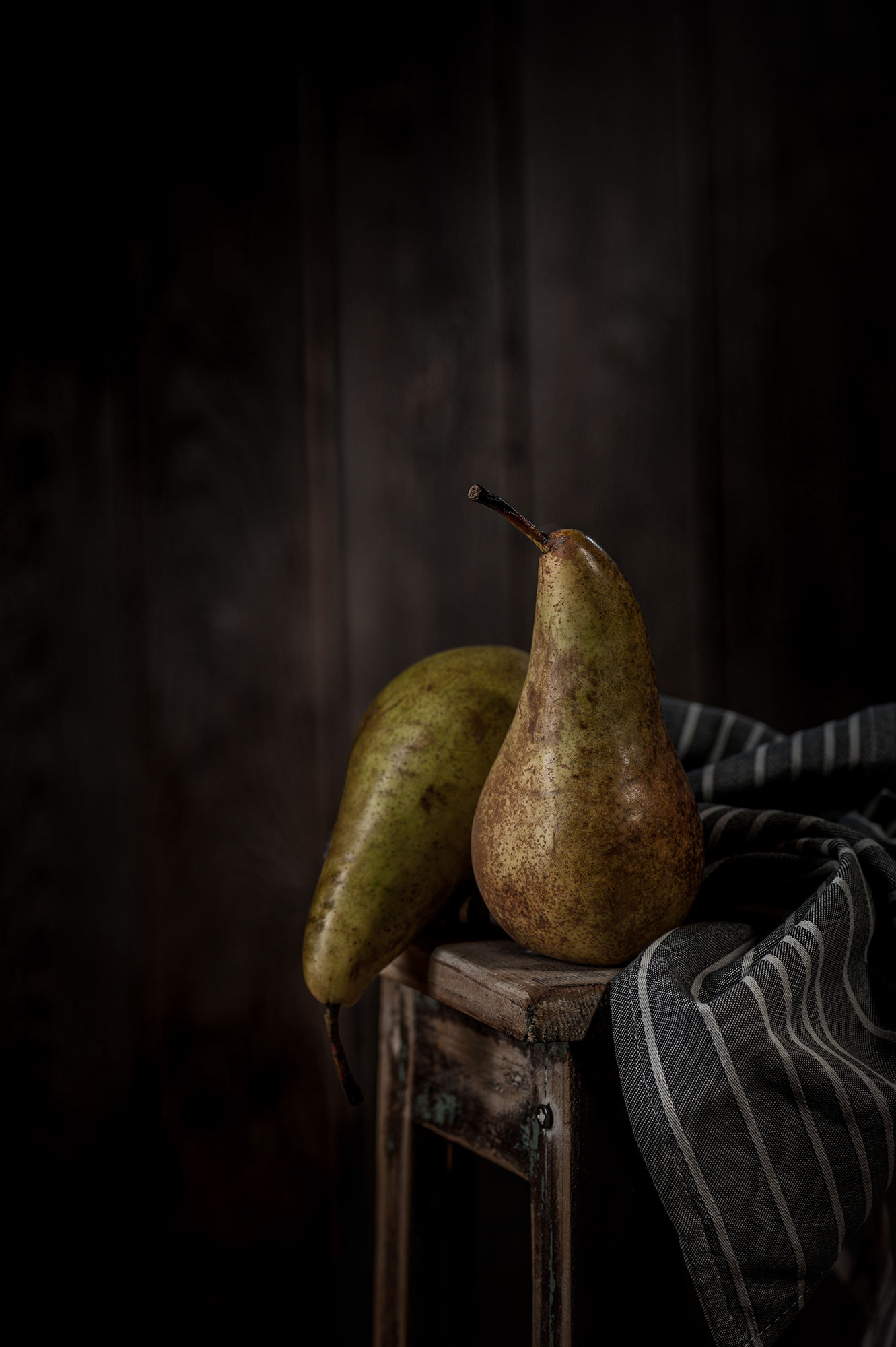 Μια υποβλητική fine art φωτογραφία νεκρής φύσης (still life) με δύο ώριμα αχλάδια πάνω σε ένα παλιό ξύλινο σκαμνί, πλαισιωμένα από μια ριγέ πετσέτα με Chiaroscuro φωτισμό, από τη φωτογράφο Βούλα Γκώτη.  An evocative fine art still life photograph featuring two ripe pears on an aged wooden stool, draped with a striped kitchen towel under moody Chiaroscuro lighting, by photographer Voula Gkoti.