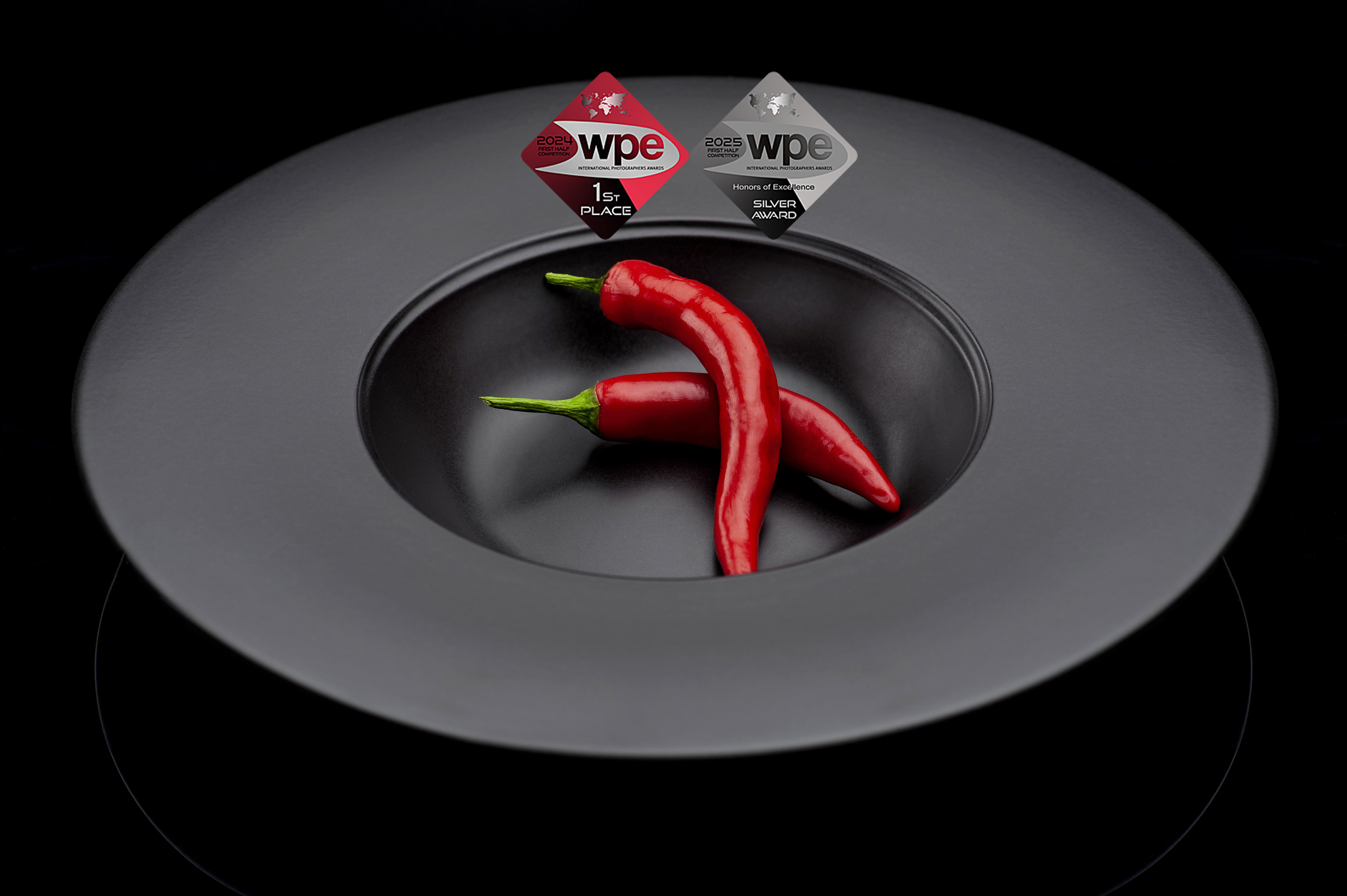 Πιάτο μαύρο με δύο κόκκινες τσίλι πιπεριές  σε μαυρο φόντο  μπαχαρικά Papadimitriou σε επαγγελματική φωτογράφιση,  chilli peppers on black  plate photography silver award photography