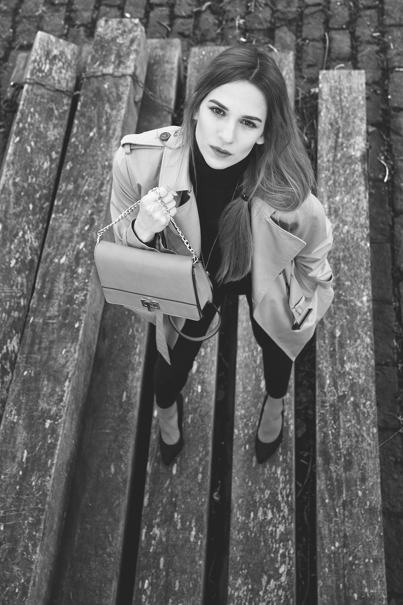 Γυναίκα με μπεζ καμπαρντίνα και τσάντα ποζάρει κοιτάζοντας προς τα πάνω, σε ξύλινη επιφάνεια.      Woman in a beige trench coat holding a handbag, looking upwards on a wooden platform.