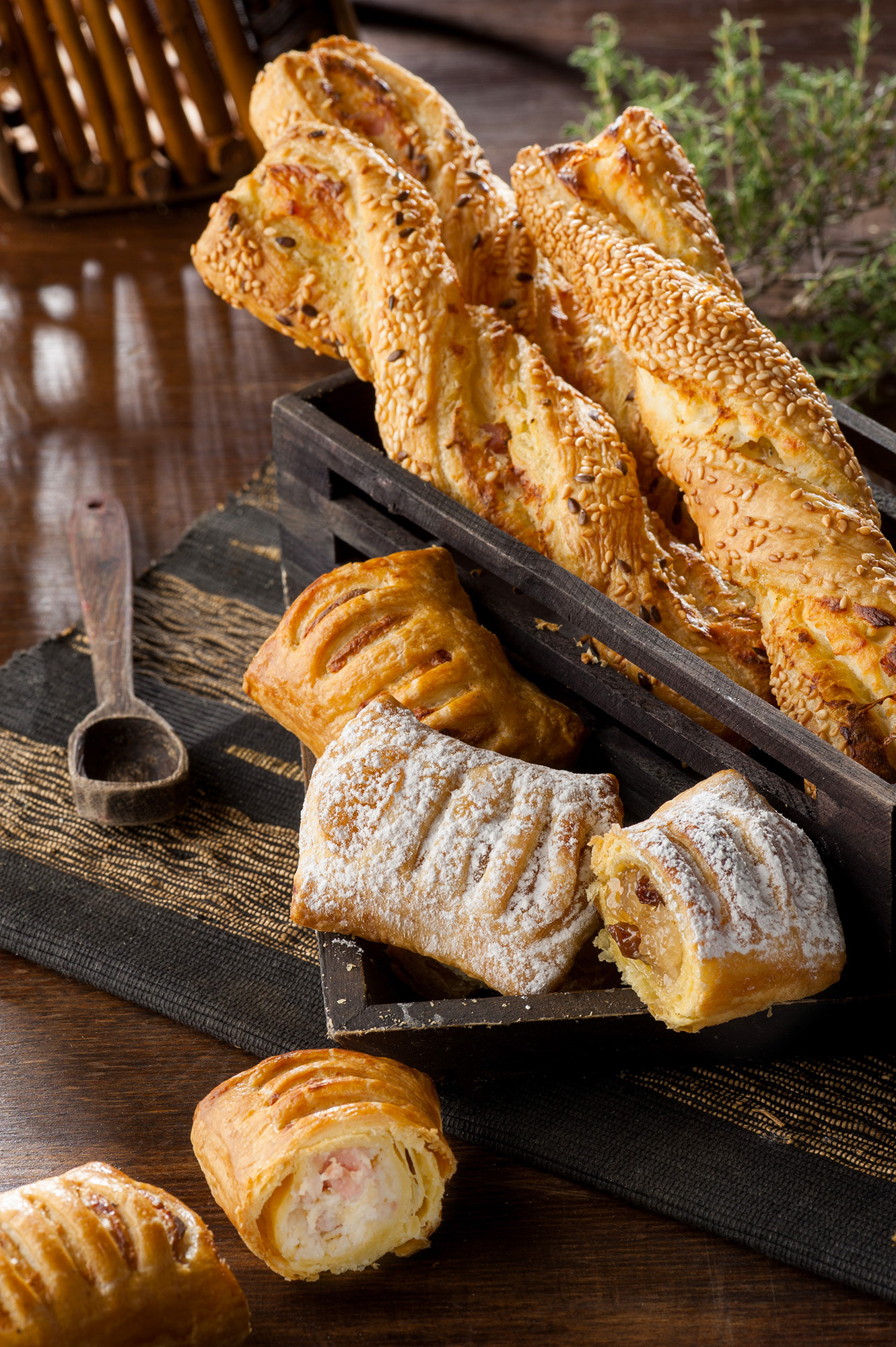 Εμπορικό food styling με χρυσαφένια στριφτάρια σφολιάτας με τυρί μέσα σε σκούρο ξύλινο κουτί, μαζί με μίνι μηλοπιτάκια με ζάχαρη άχνη και αλμυρά ρολά με ζαμπόν και κασέρι σε ρουστίκ φόντο.   /  Commercial food styling of golden puff pastry cheese twists in a dark wooden box, paired with powdered sugar mini apple pies and savory ham and cheese rolls on a rustic background.