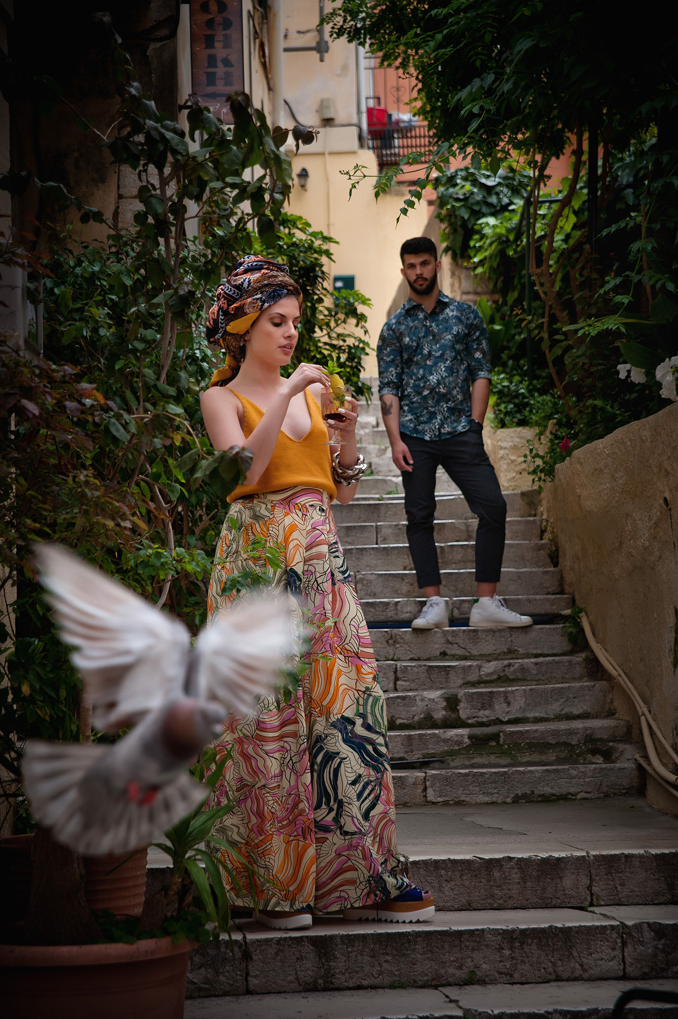 Γυναίκα με πολύχρωμο ρούχο κατεβαίνει σκαλιά της πόλης ενώ ένα περιστέρι πετάει στο προσκήνιο και ένας άνδρας φαίνεται στο βάθος.,     Woman in a vibrant outfit walking down stone stairs, with a man in the background and a bird flying across the frame.    