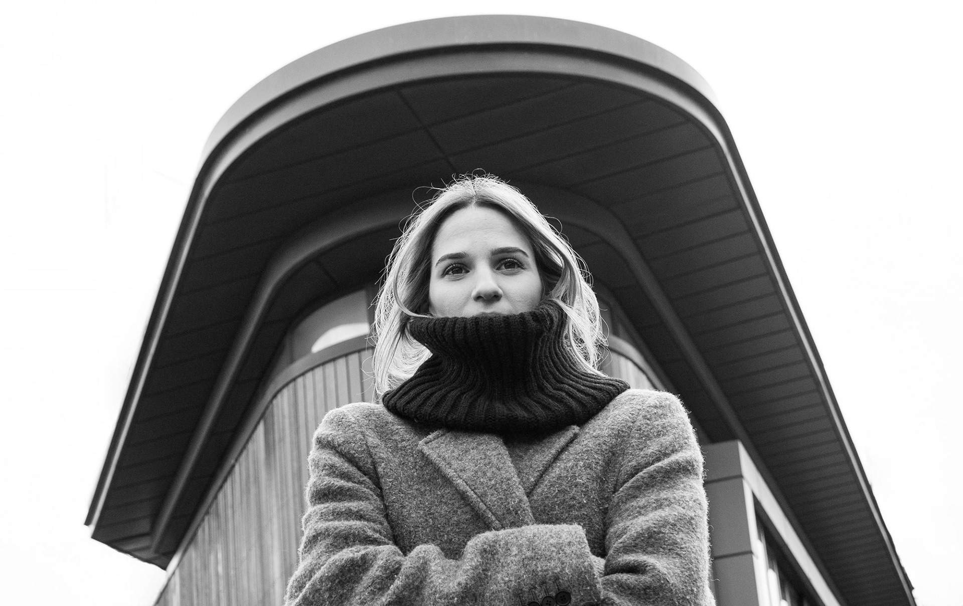 Πορτραίτο γυναίκας με μάλλινο παλτό και ψηλό ζιβάγκο, μπροστά από αρχιτεκτονικό φόντο.    Portrait of a woman in a wool coat with a high turtleneck, standing against an architectural backdrop.