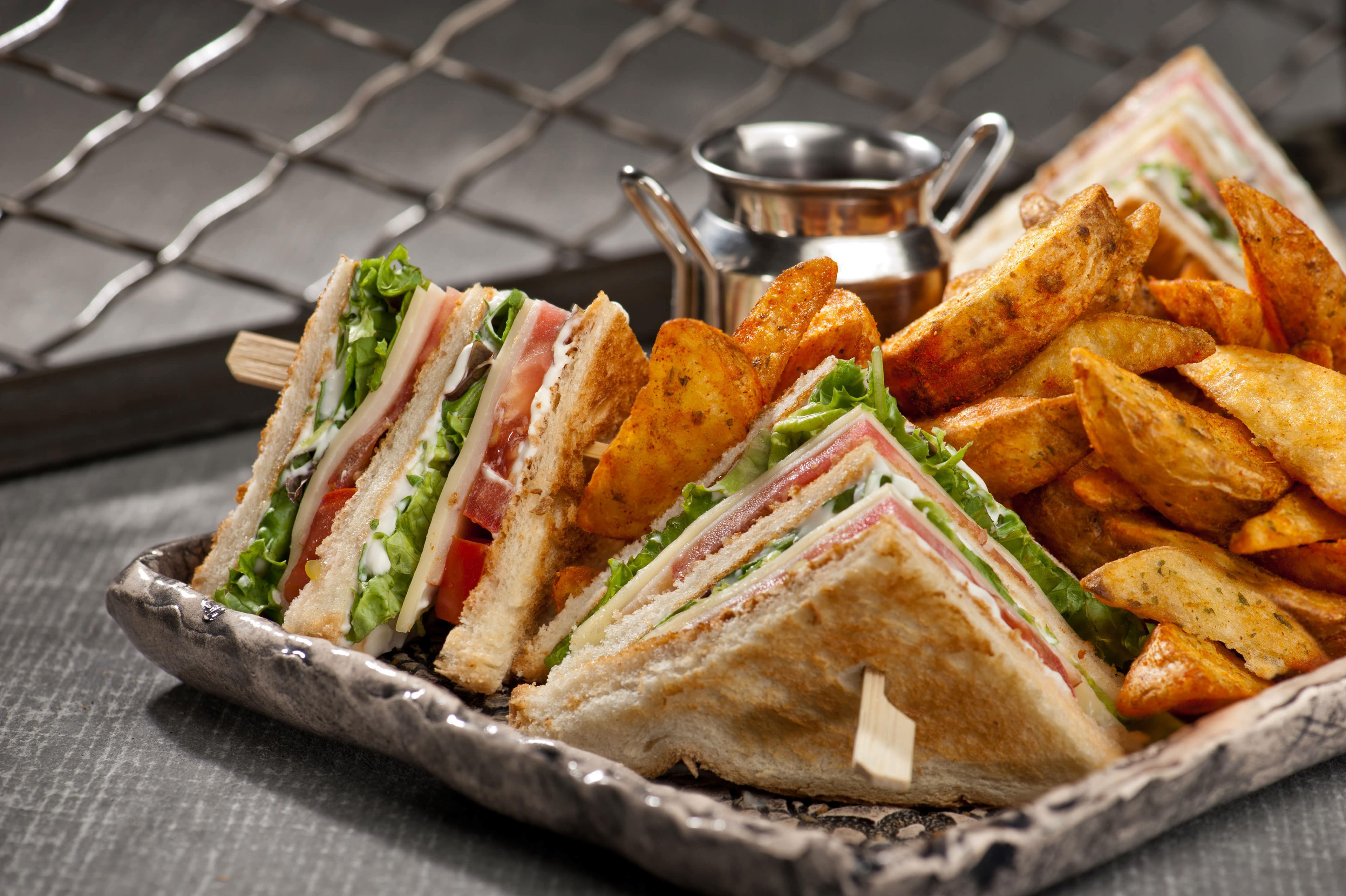 φωτογράφιση club sandwich σε πιάτο με πατατες καντρυ , ντομάτα, σως τυριού, κασέρι πάνω σε γκρί τραπεζι, photography of a club sandwich on a plate with country potatoes, tomato, cheese sauce, and cheese on a grey table
