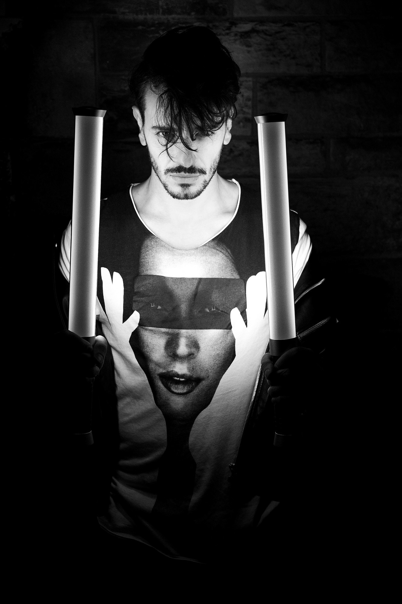 Άνδρας με μπλουζάκι γραφιστικού σχεδίου και έντονο φωτισμό κρατά δύο κάθετες λάμπες στο σκοτάδι.     Man wearing a graphic T-shirt holding two vertical lights in a dark, moody scene.