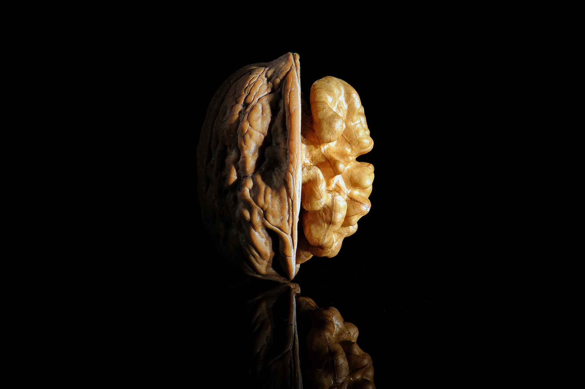 Μια μινιμαλιστική macro φωτογραφία food art που αποτυπώνει ένα μισό καρύδι να ισορροπεί όρθιο πάνω σε μια μαύρη, γυαλιστερή επιφάνεια με καθαρή είδωλη αντανάκλαση, από τη φωτογράφο Βούλα Γκώτη.  A minimalist macro food art photograph capturing a halved walnut shell balanced upright on a glossy black surface with a crisp mirror reflection, by photographer Voula Gkoti.