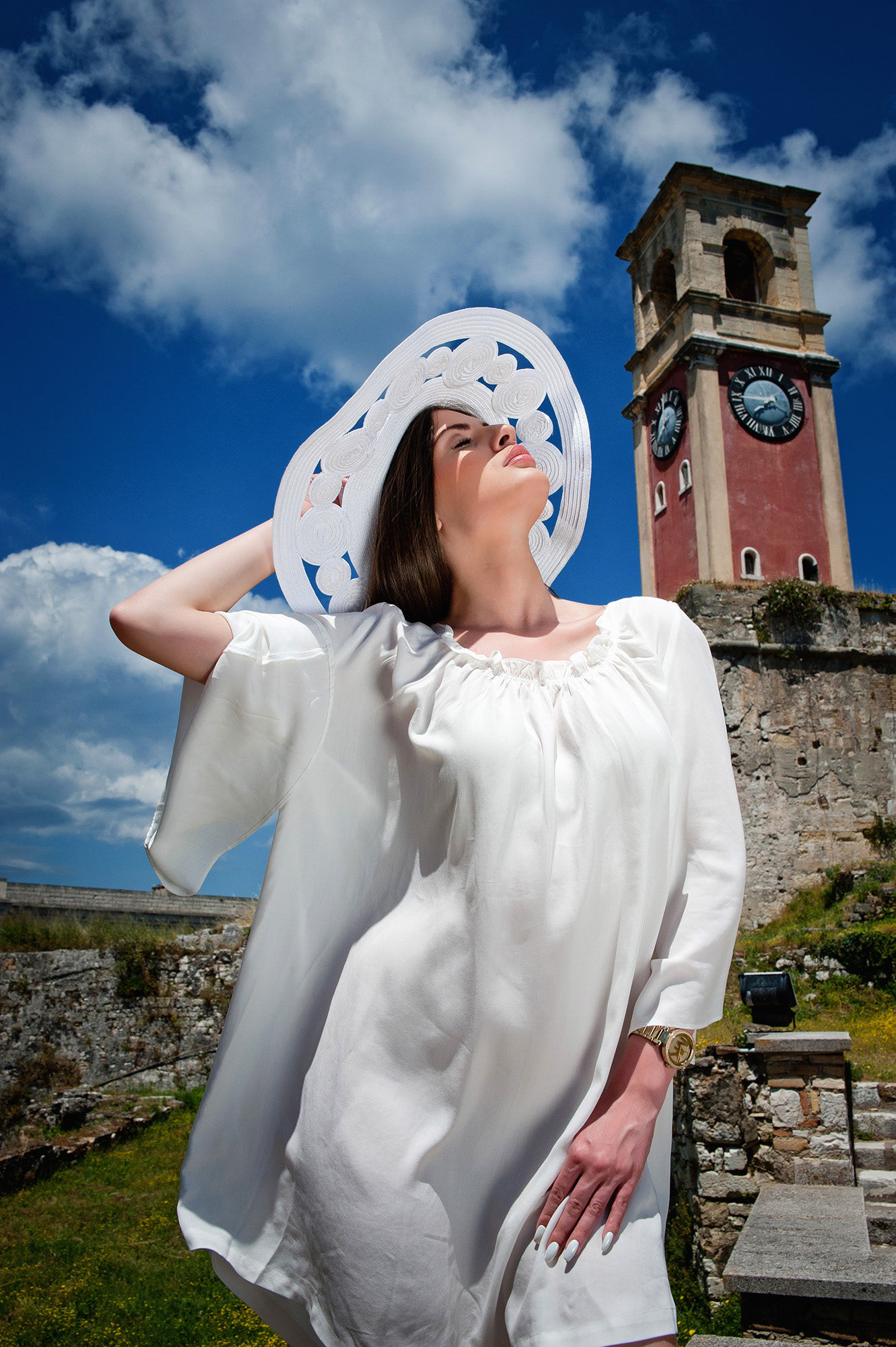 Γυναίκα με λευκό φόρεμα και μεγάλο καπέλο απολαμβάνει τον ήλιο μπροστά από ρολόι-πύργο.,    Fashion portrait of a woman in a white dress and wide hat under bright sunlight with a clock tower in the background.,