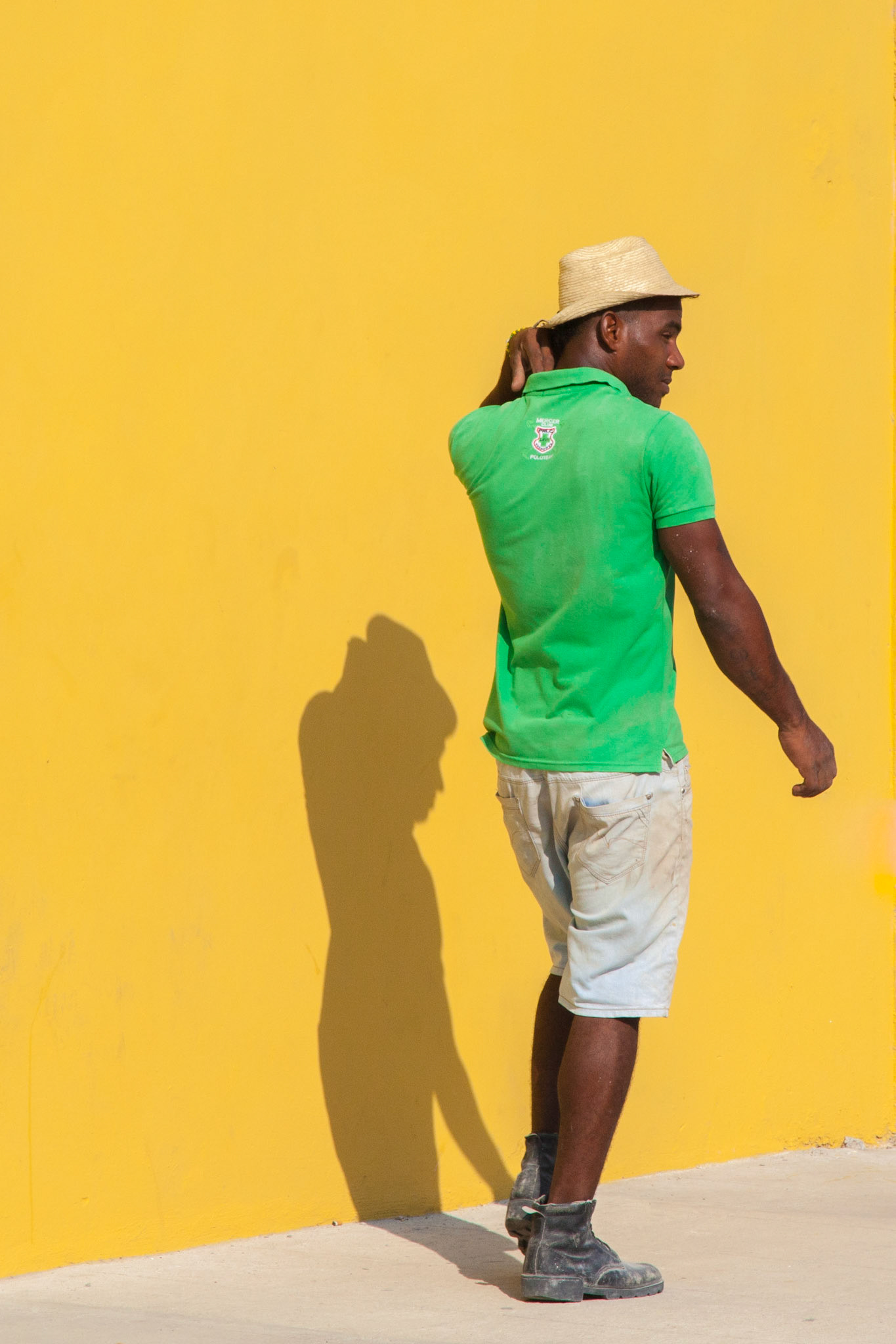 A construction worker in Havana walks along a sunlit sidewalk, his vibrant attire complementing the city’s famously colorful streets. Shadows and sunlight create a spontaneous portrait of everyday Cuban life.
