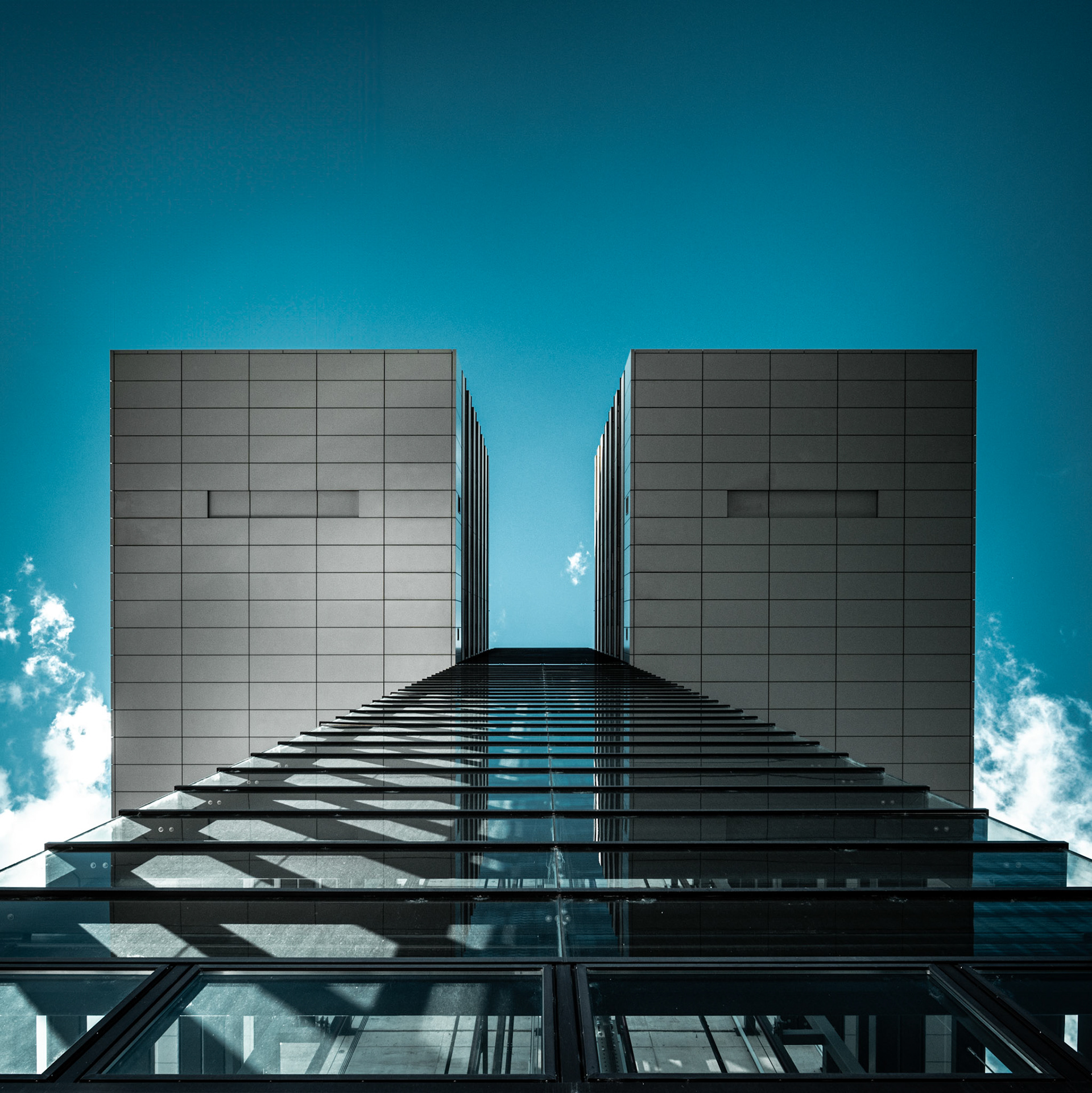 The Kranhäuser stand tall against a vibrant blue sky, their distinctive top-heavy architecture mimicking the form of dockside cranes. Symmetrical staircases lead the eye upwards to where the two structures meet, framing a slice of the open sky and a wispy cloud, blurring the lines between urban innovation and the natural canvas above.