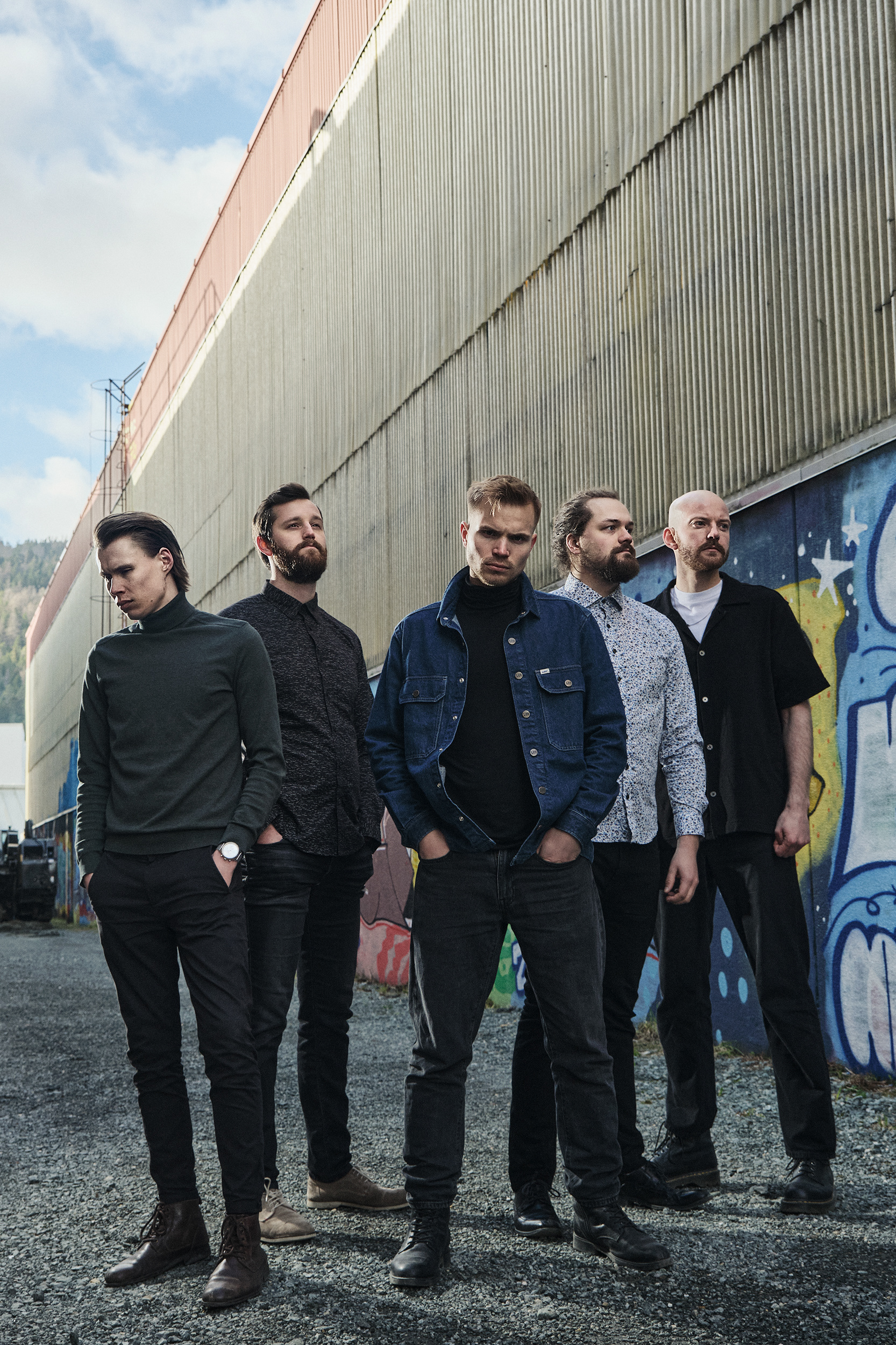 Gruppeportrett av fem bandmedlemmer i et røft, industrielt miljø med graffitivegger og dramatisk, hard lyssetting. Bildet brukes som del av bandets visuelle profil og formidler en rå og urban stemning.