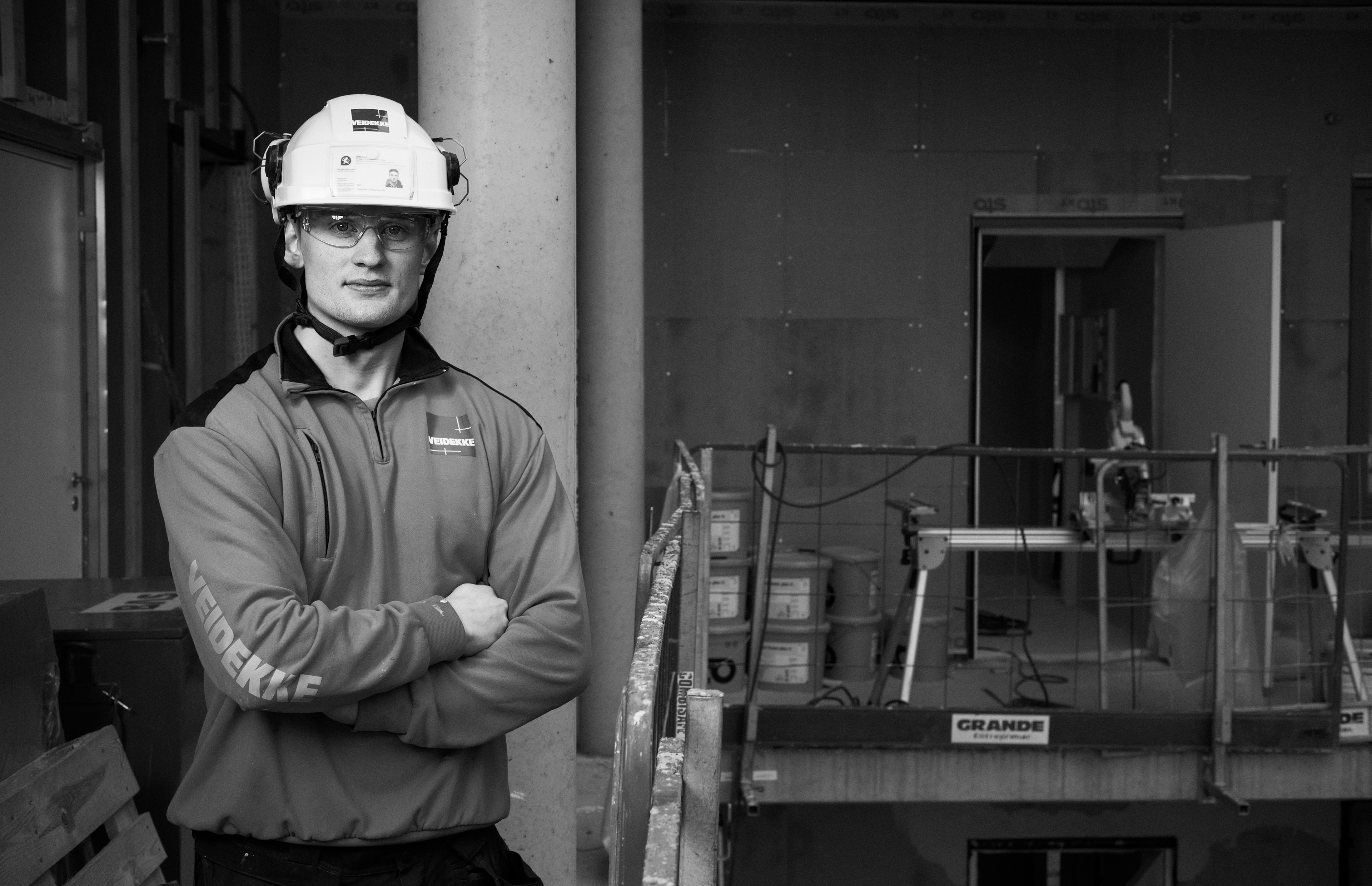 Portrett av mann i sikkerhetshjelm og arbeidsklær på byggeplass omgitt av byggematerialer. Han står posert med et vennlig og selvsikkert uttrykk. Bildet viser medarbeideren i sitt miljø for å bygge tillit til bedriften.