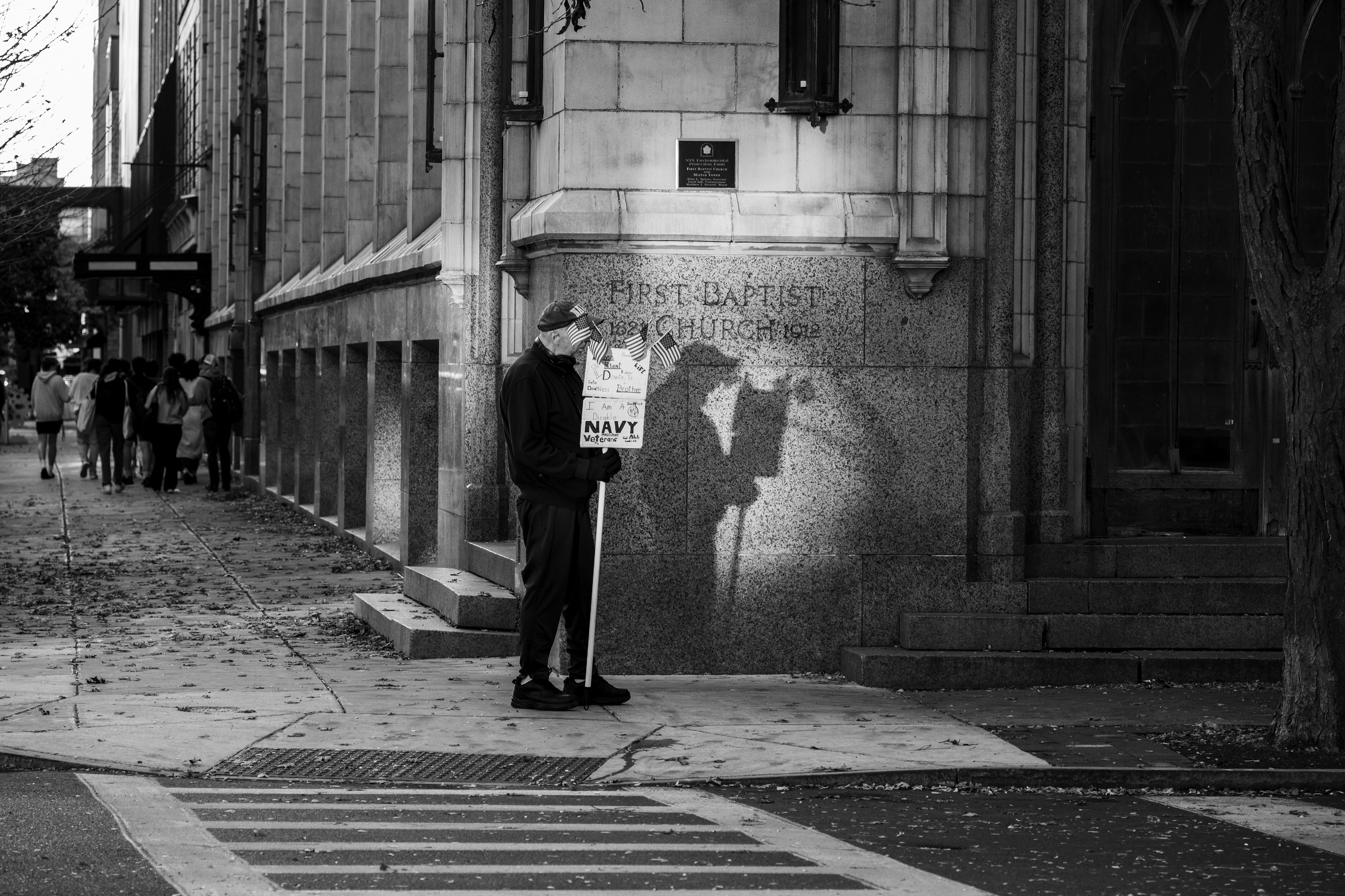 A disabled Navy veteran stands alone on a city sidewalk in front of the First Baptist Church in Syracuse, NY, holding a handmade sign that reflects his service and sacrifice. Like many retired veterans, he faces challenges related to disabilities or health conditions acquired during or after his service.