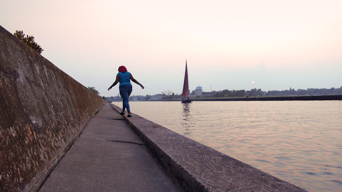 Woman walking along the canal in Kenosha Wisconsin with the Red Witch 2 sailboat in the distance.