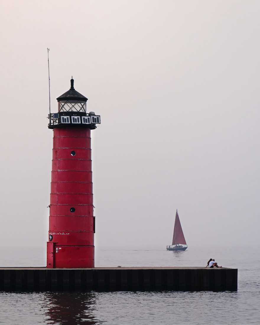 Kenosha North Pier Lighthouse with the iconic Red Witch 2 sailboat in the background and a couple sitting on the pier.