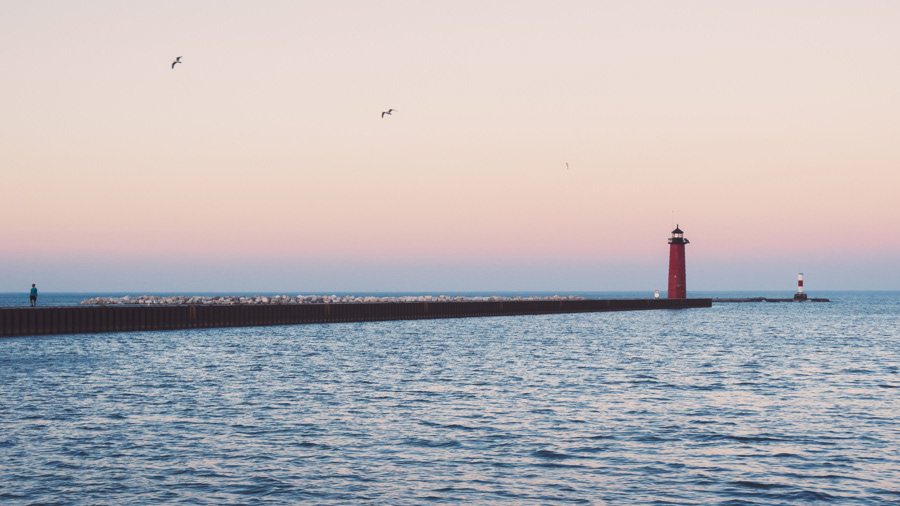 The pier of the red Kenosha Lighthouse at sunset.  Three birds fly away from the lighthouse towards a man at the end of the pier.