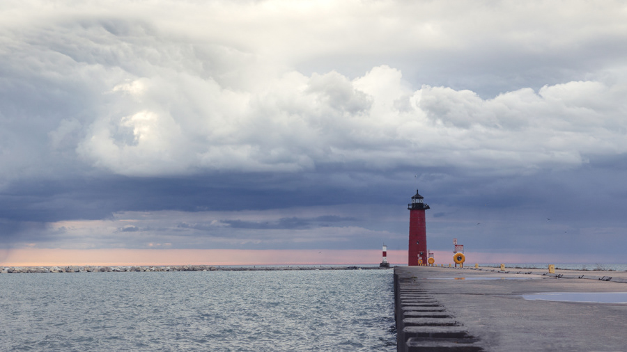 A moody sky with dark clouds swirling over Lake Michigan and the pier of the red Kenosha Lighthouse on Simmons Island in Kenosha Wisconsin.